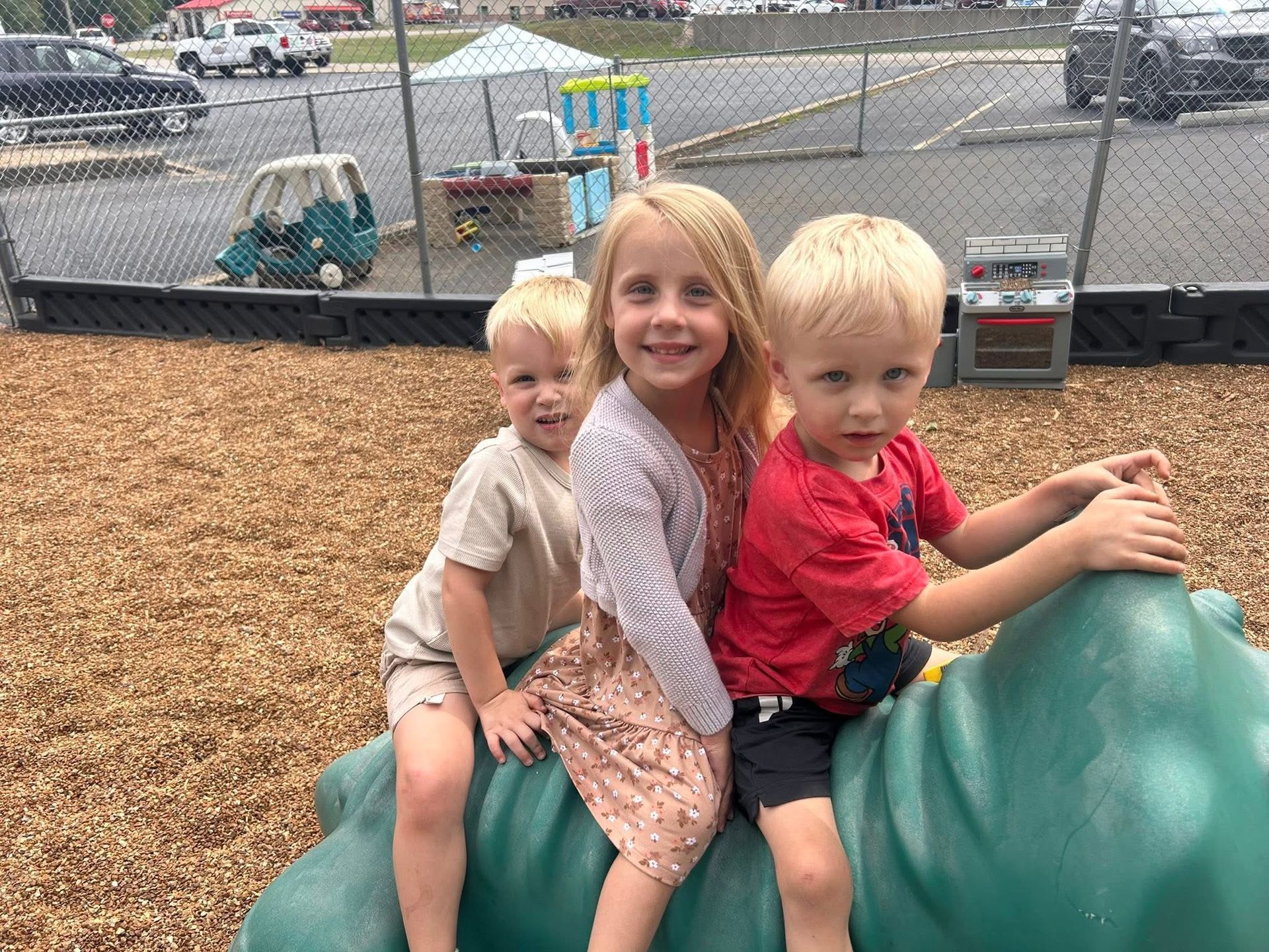 Three children on a green play structure, smiling. Playground setting.