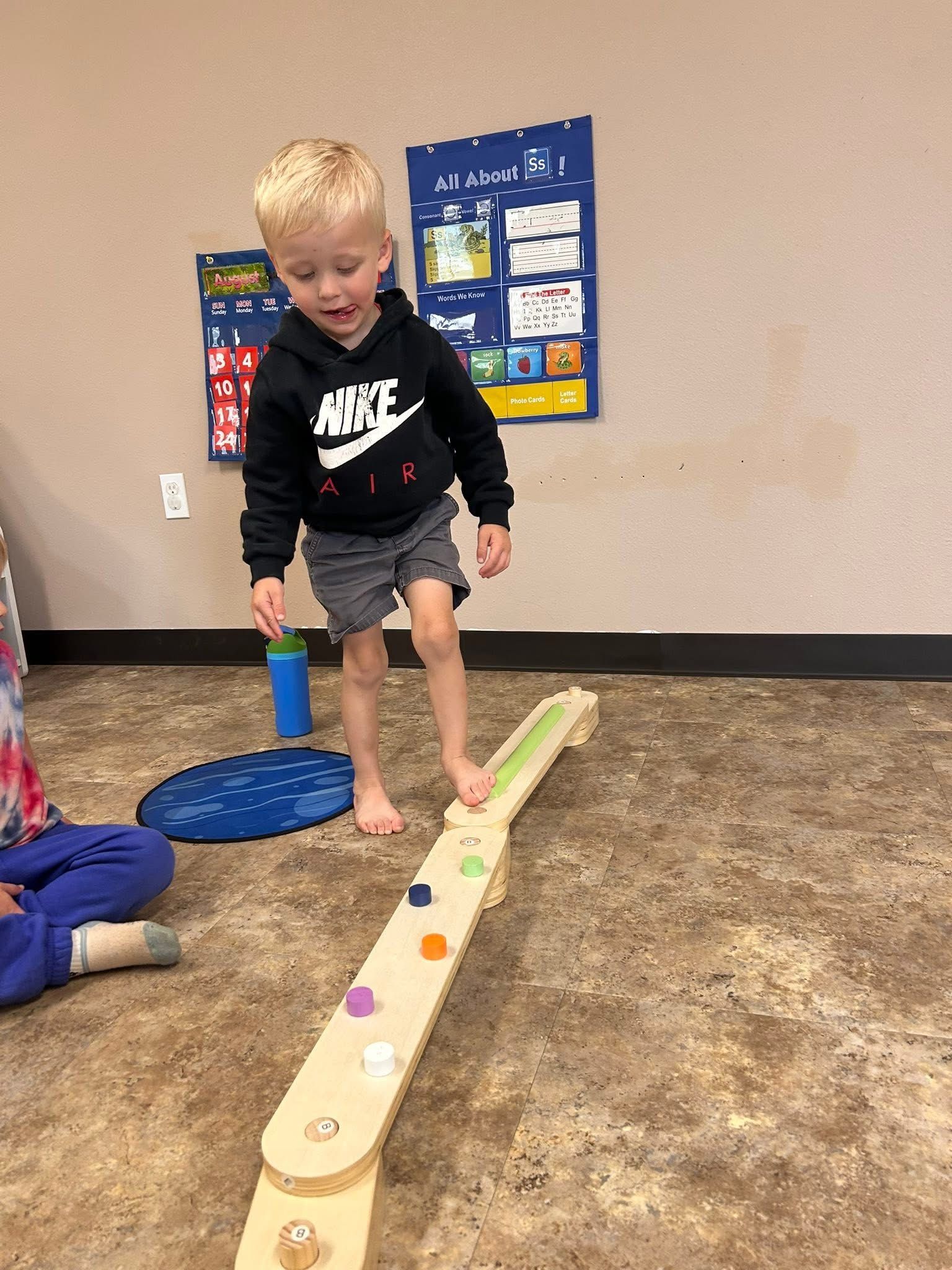 Young child wearing a Nike hoodie balances on a wooden seesaw, eyes focused, indoors.