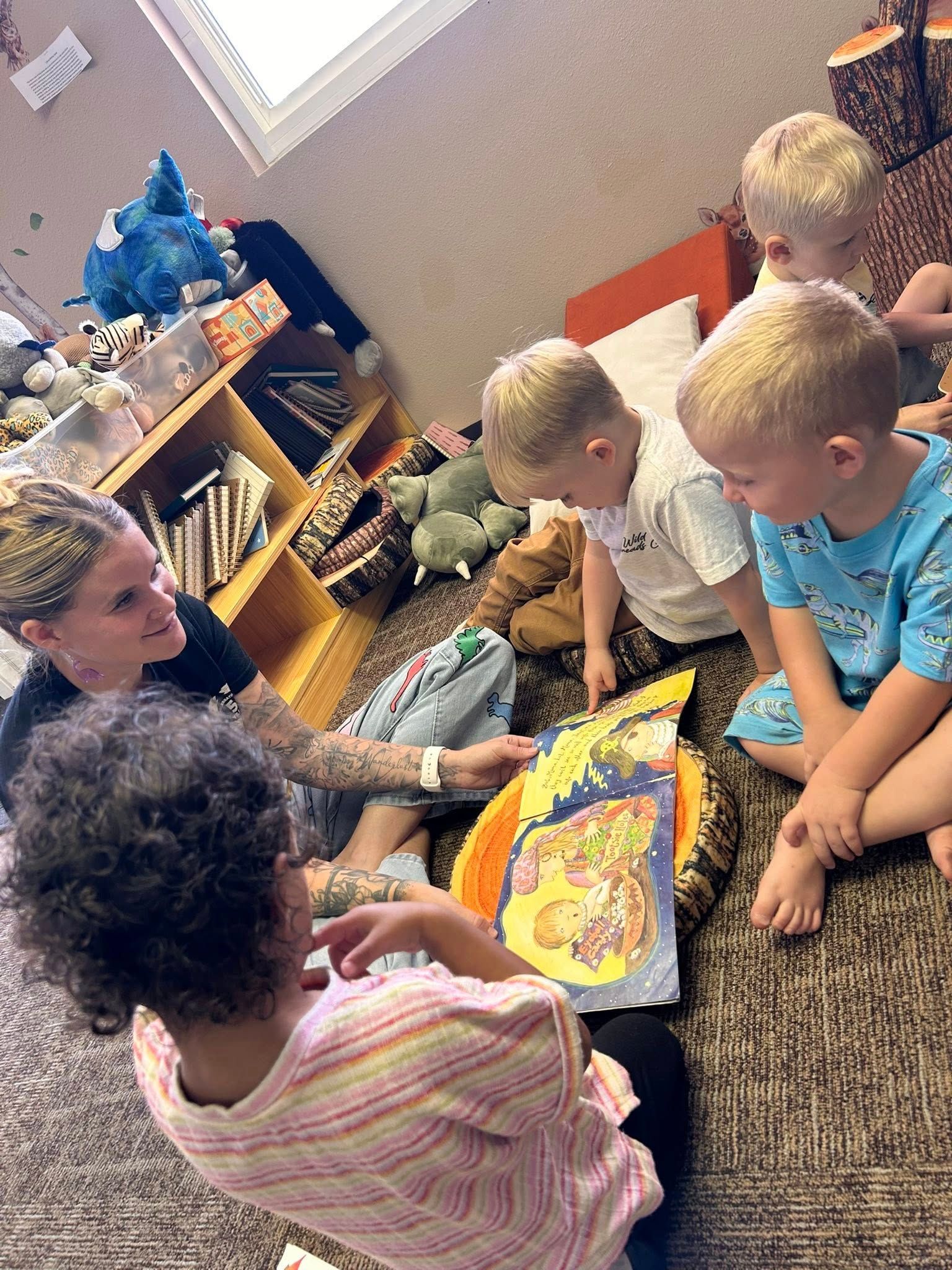 A woman reads a book to four children sitting on a carpeted floor.