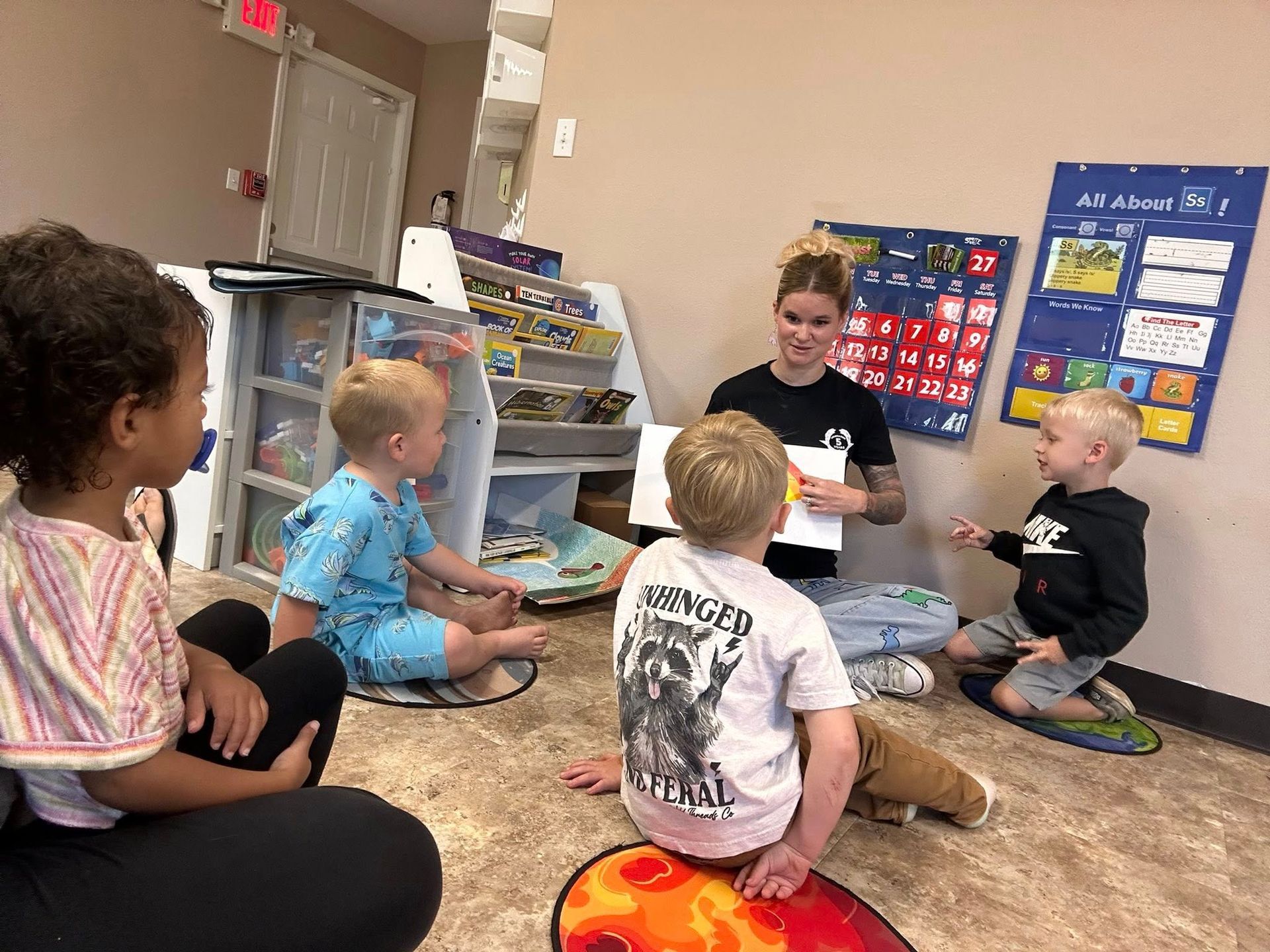 Teacher reading to children on floor. Room has books and calendar.