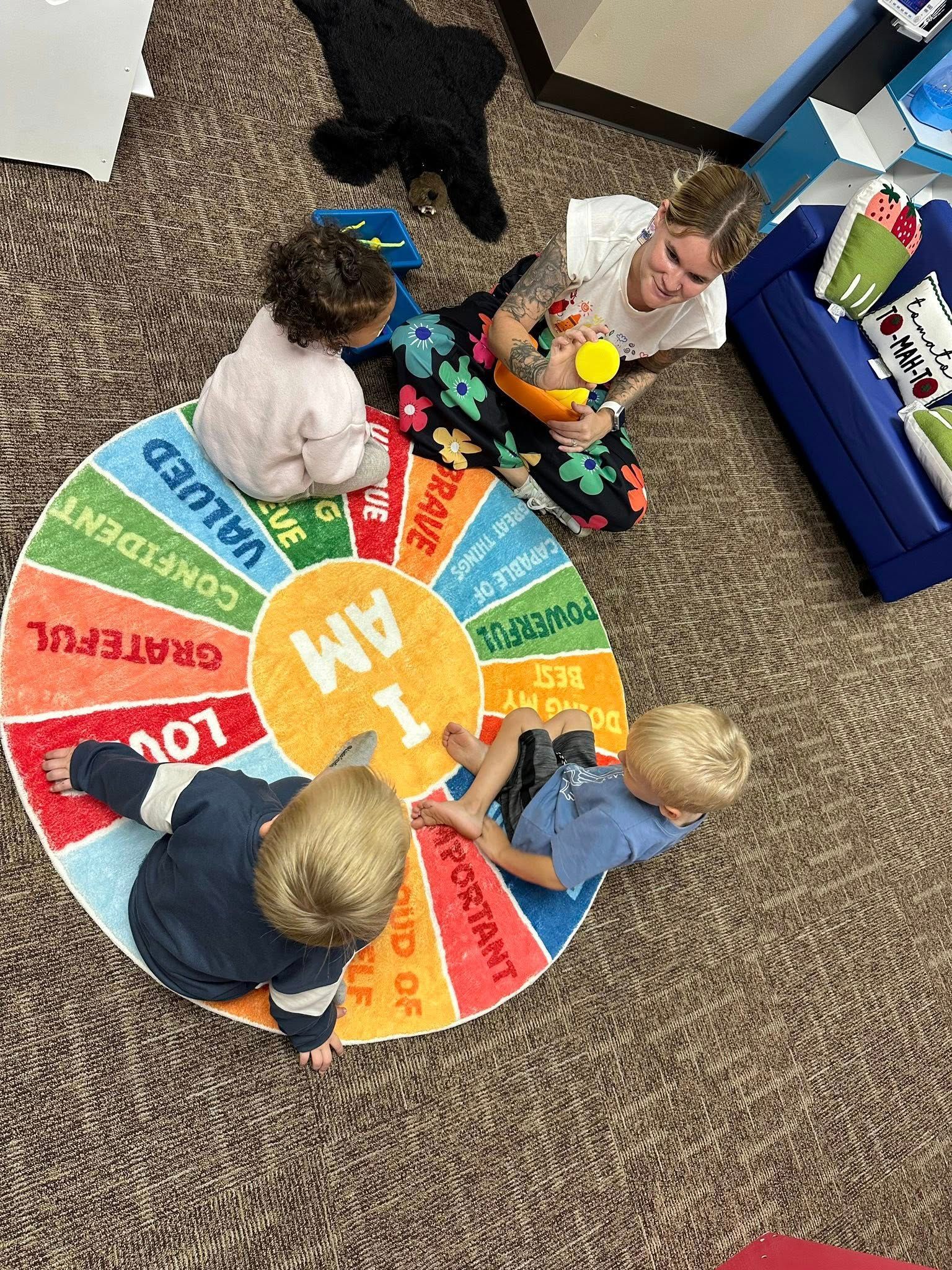 Children and adult playing with a “I AM” wheel rug. The rug has positive affirmations. Indoors.