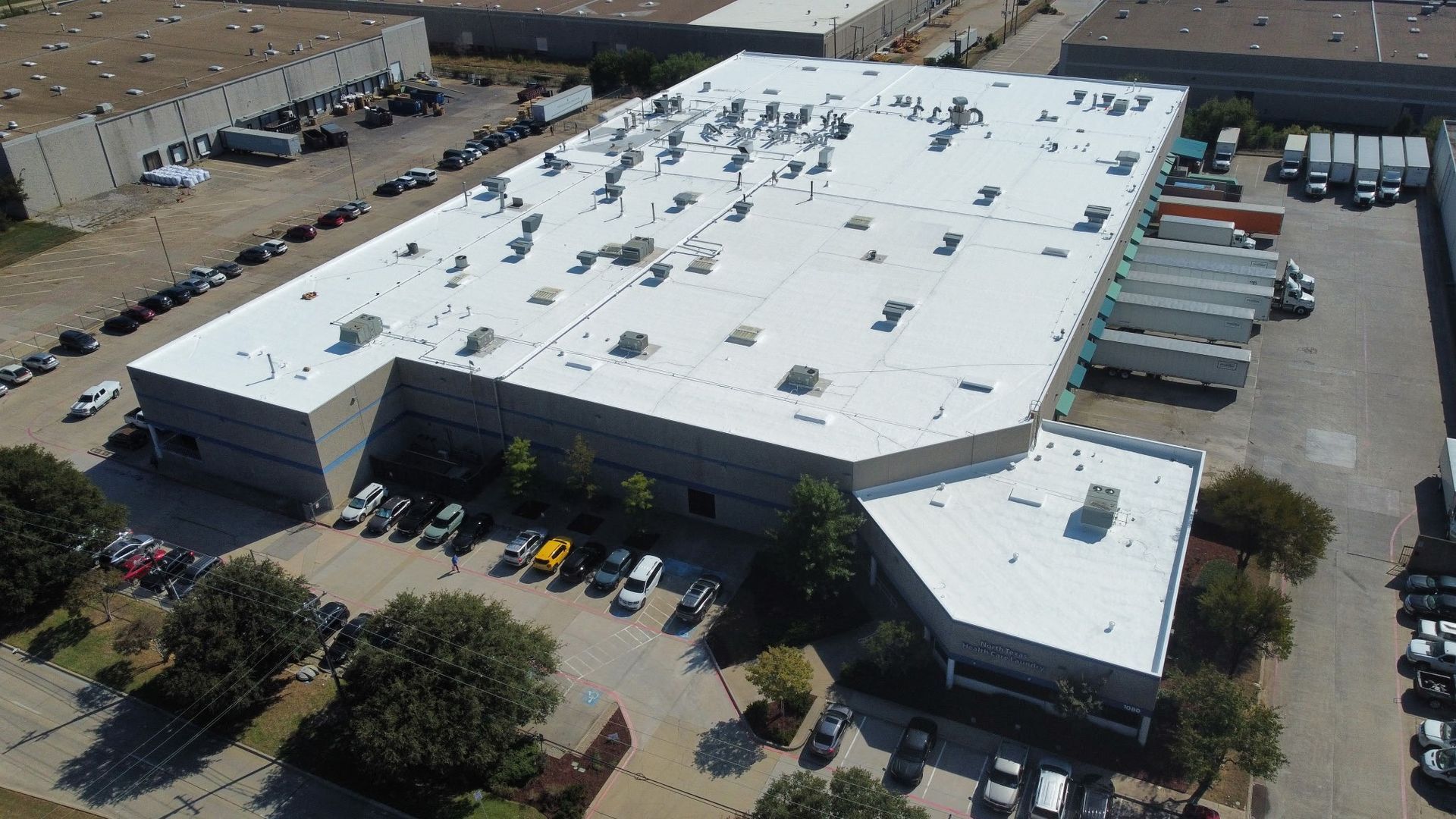 Aerial view of a large white-roofed commercial building with parked cars and trucks in a lot