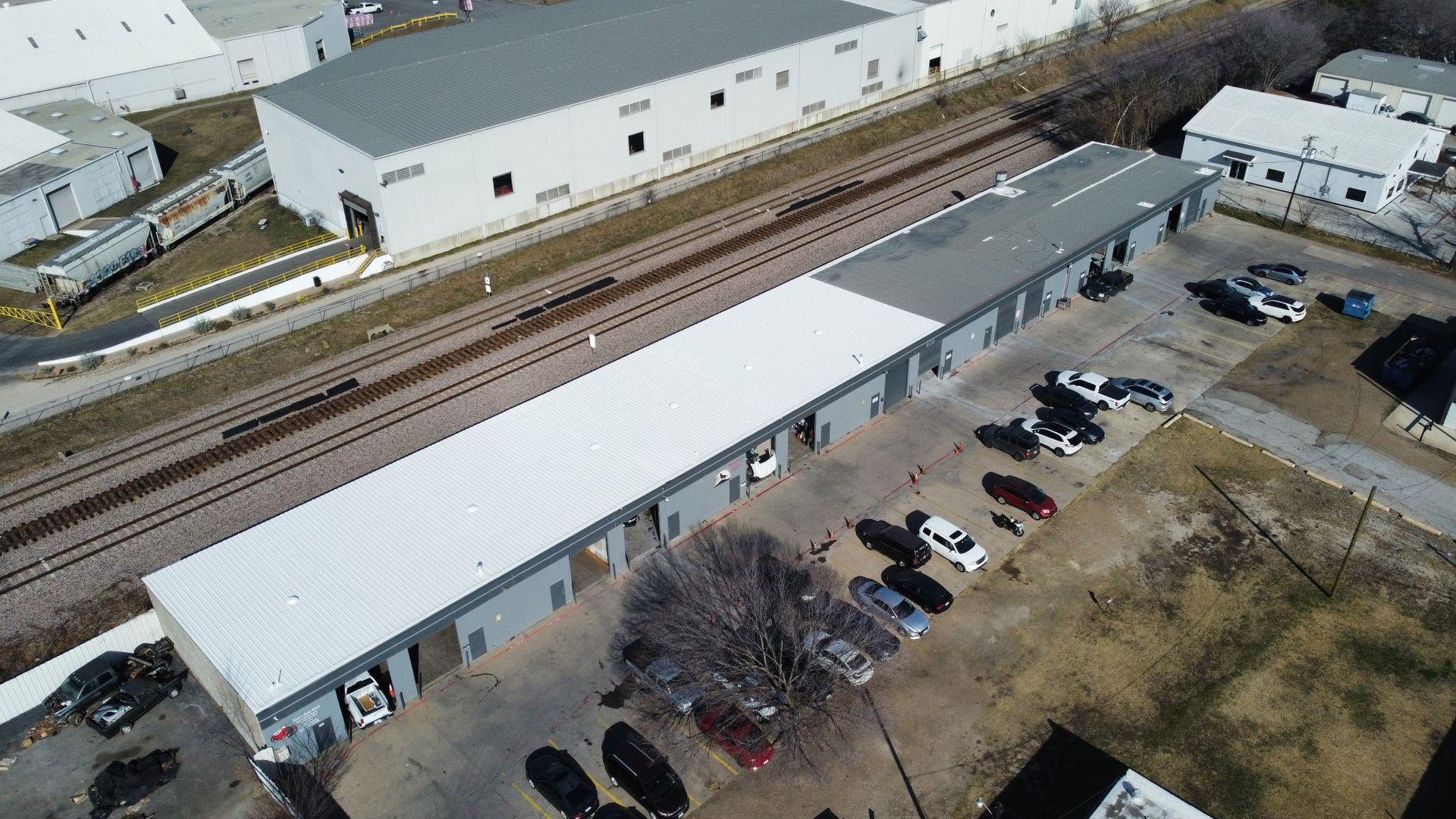 Aerial view of a long industrial building beside railroad tracks and a parking lot with several cars.