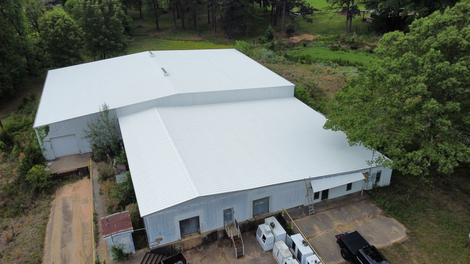 Aerial view of two large white-roofed buildings surrounded by trees and parked vehicles.
