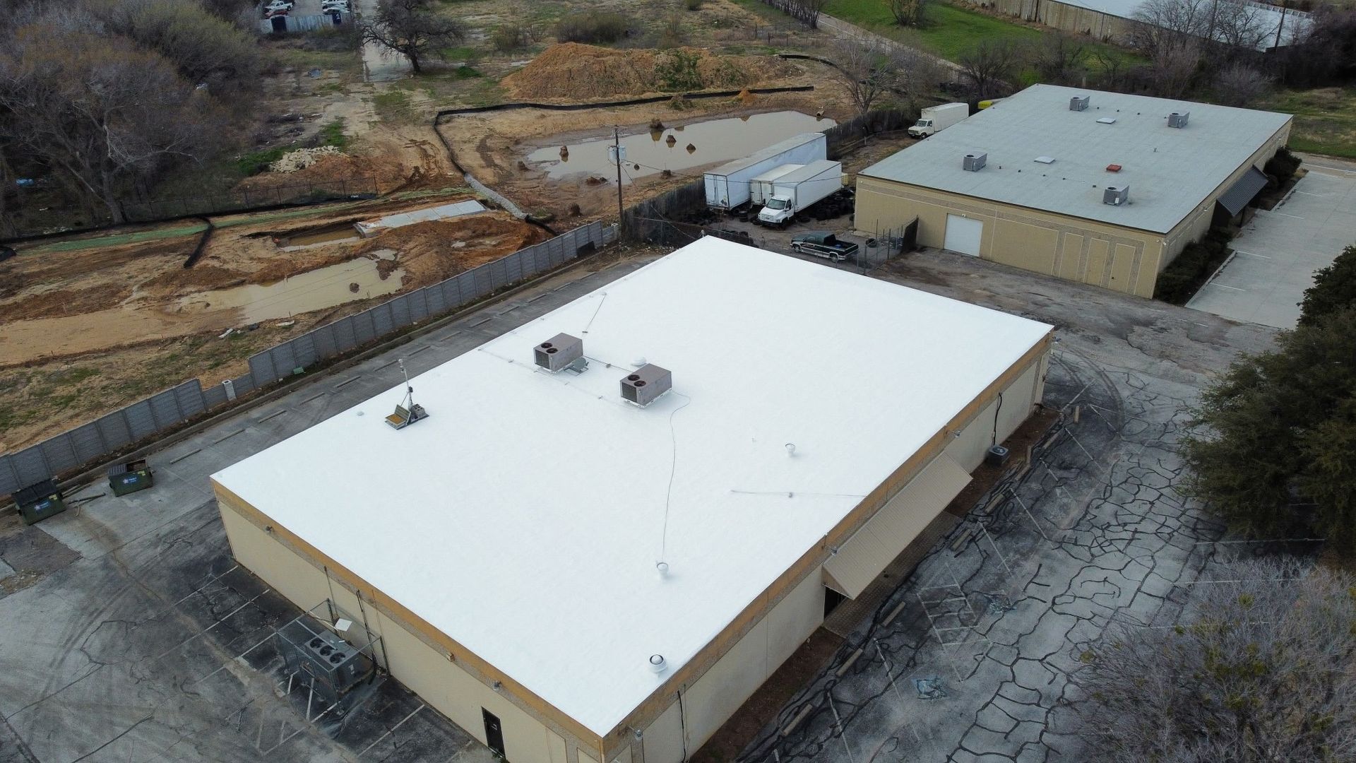 Aerial view of two beige industrial buildings with white roofs beside a paved lot and wooded hillside