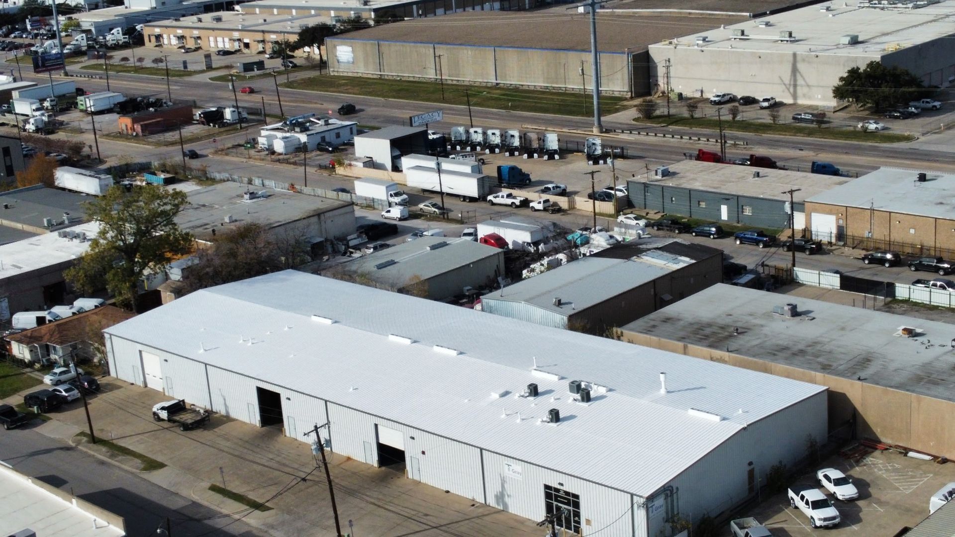 Aerial view of white industrial warehouse buildings in a commercial area with parked trucks and cars.