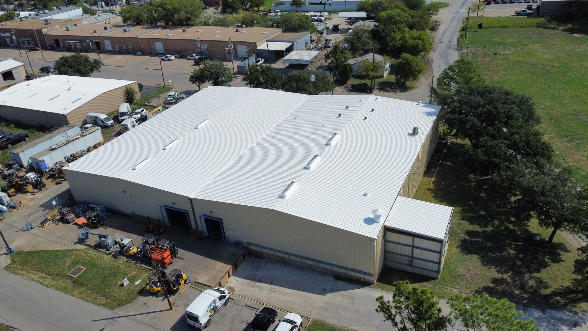 Aerial view of a large white warehouse with parked vehicles and surrounding trees and roads