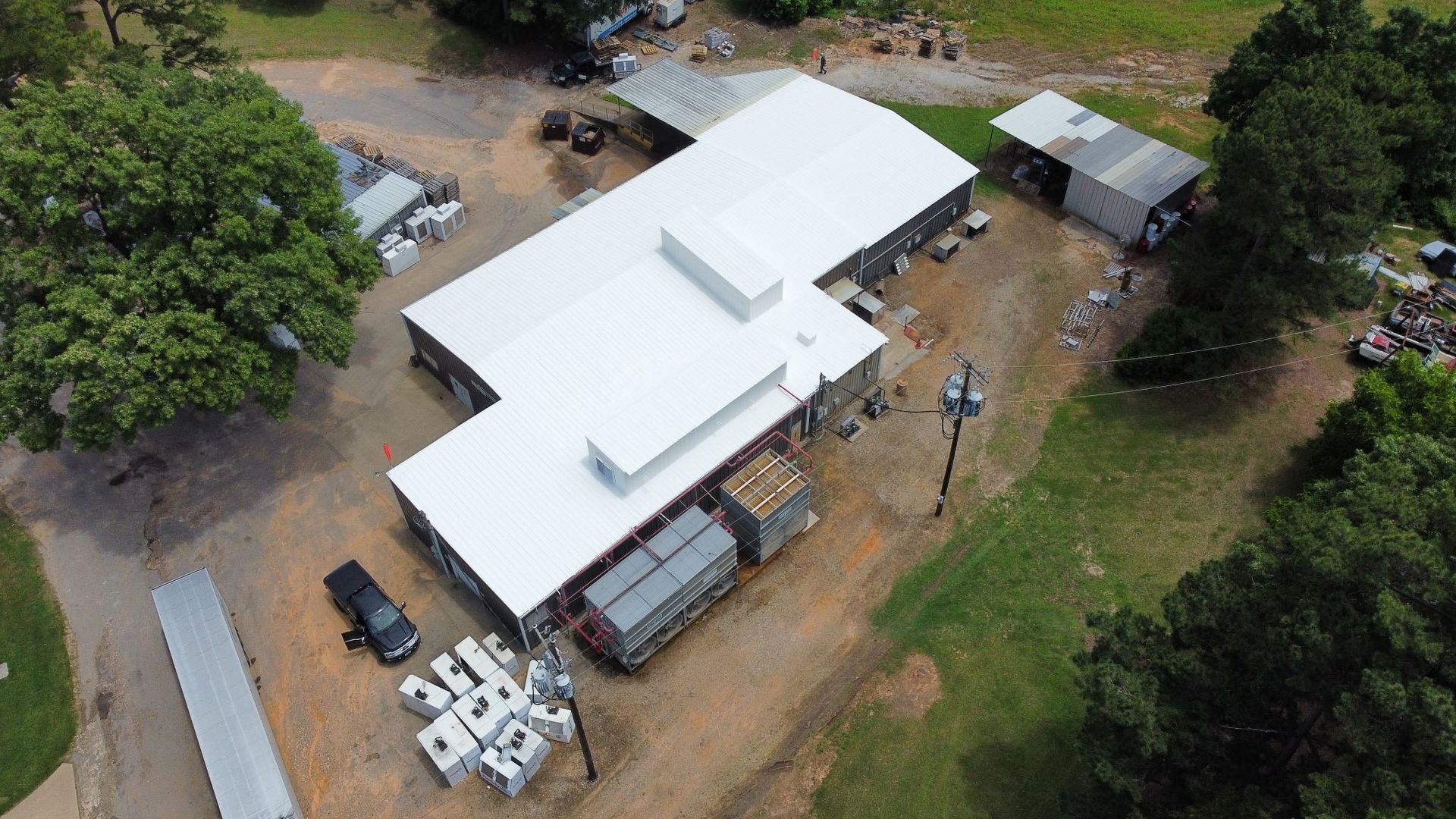 Aerial view of a white-roofed building with parked vehicles, trees, and dirt paths on rural property