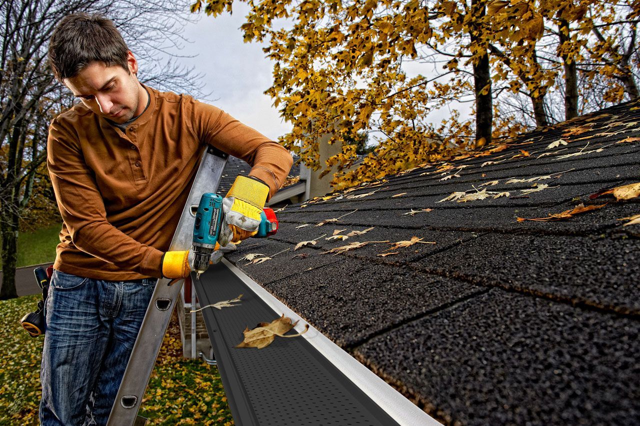 Man on ladder installing Alu-Rex gutter guard on a roof. He wears a brown shirt, gloves, and uses a drill. Autumn foliage in the background.