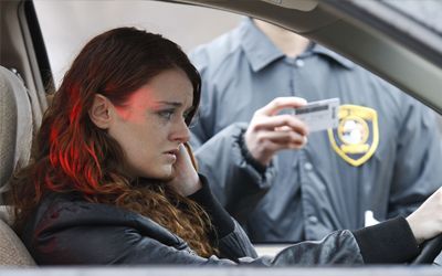 A woman is sitting in a car talking on a cell phone while a police officer takes a picture of her.