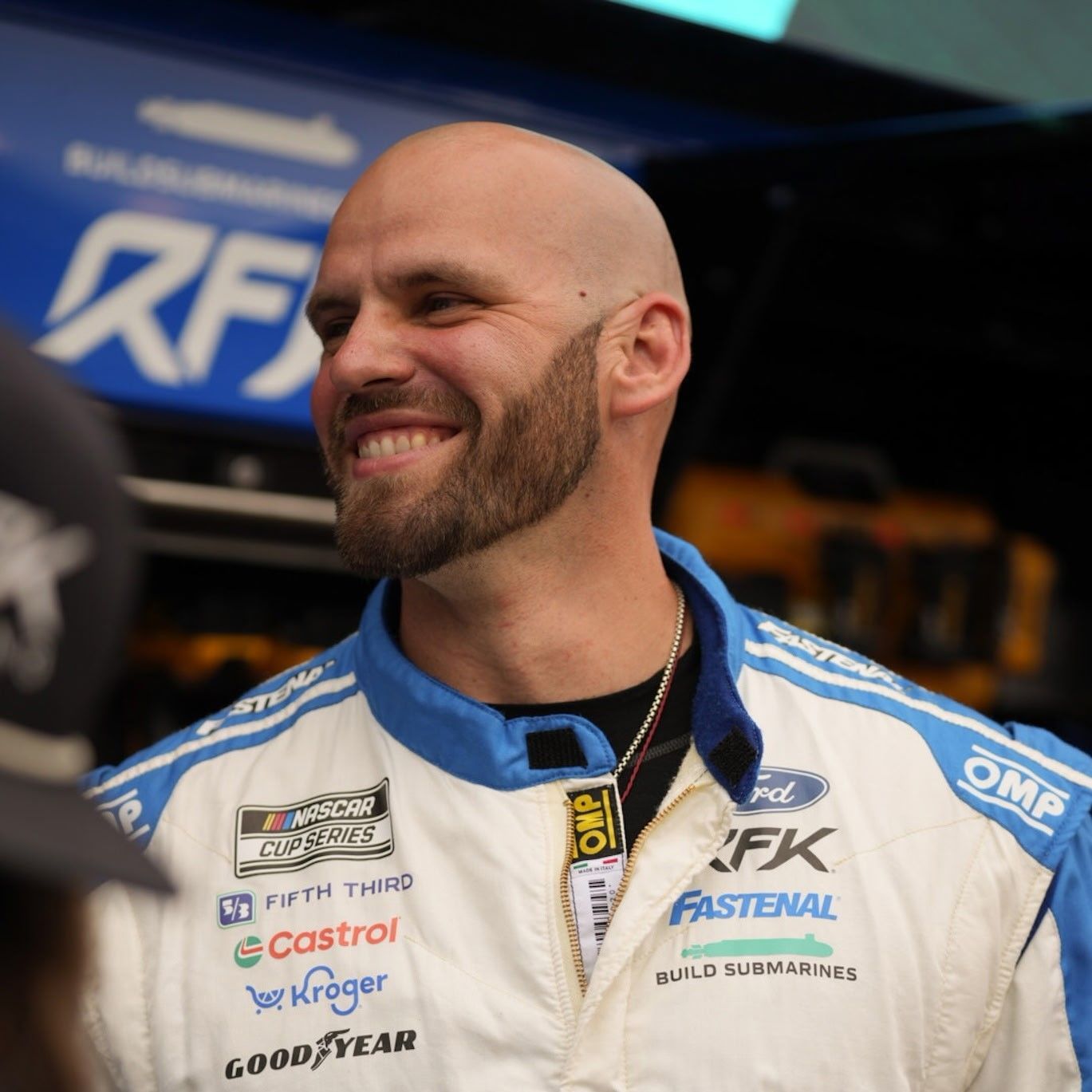 Smiling race driver in a white and blue suit standing in front of a race car garage