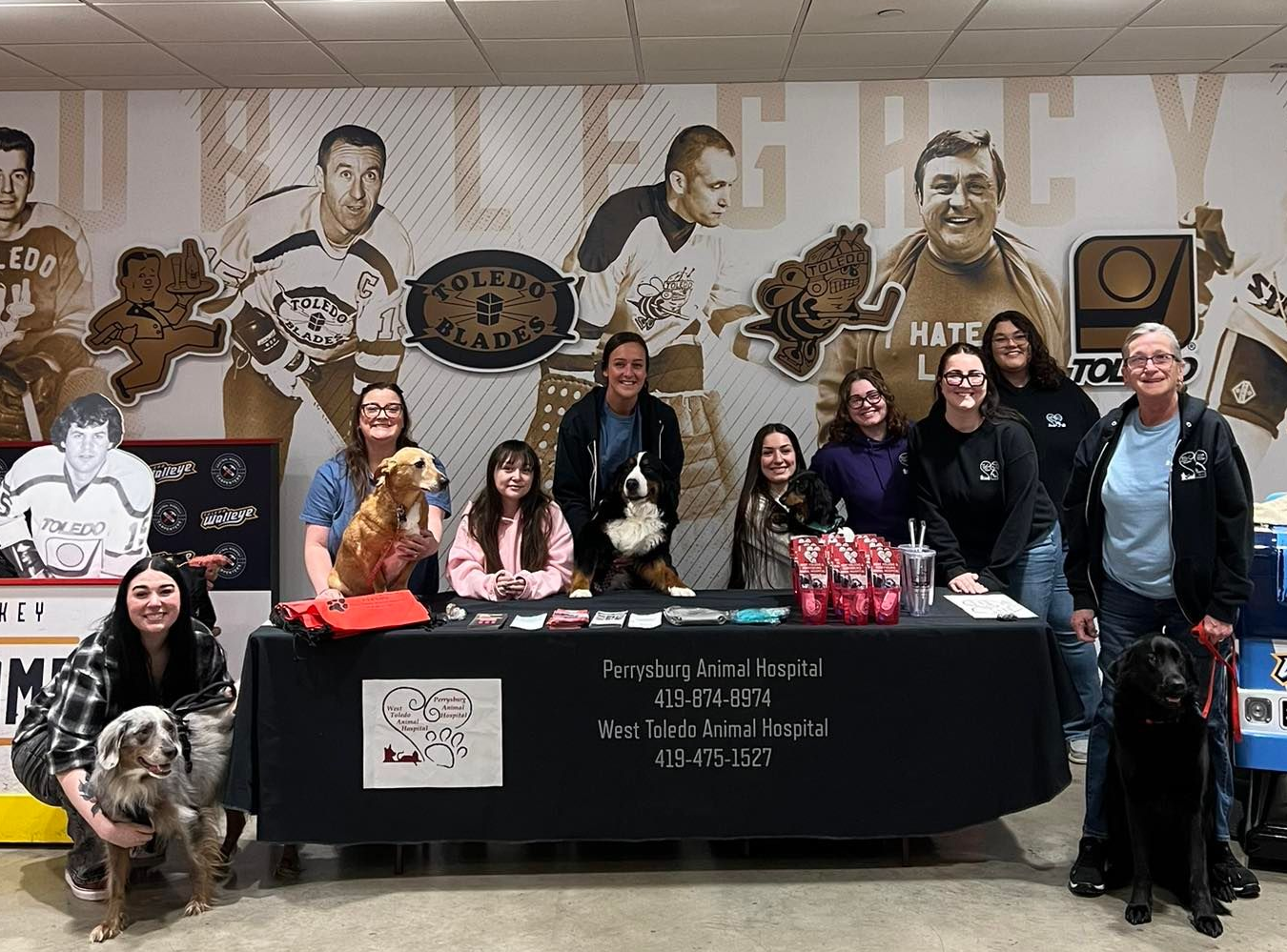 Group posed behind a black table at a pet event, with dogs, portraits on the wall, and a blue truck display.