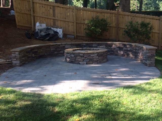 Stone fire pit and seating area in a backyard, with wooden fence backdrop and green lawn.