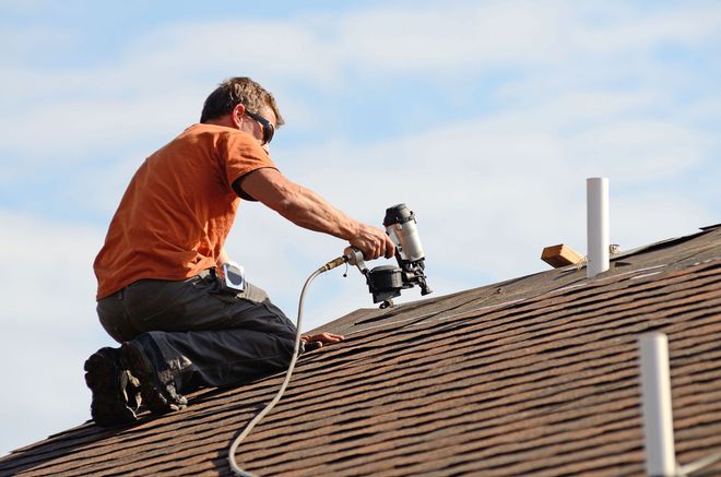 Worker using a roofing nail gun on a shingled roof under a blue sky