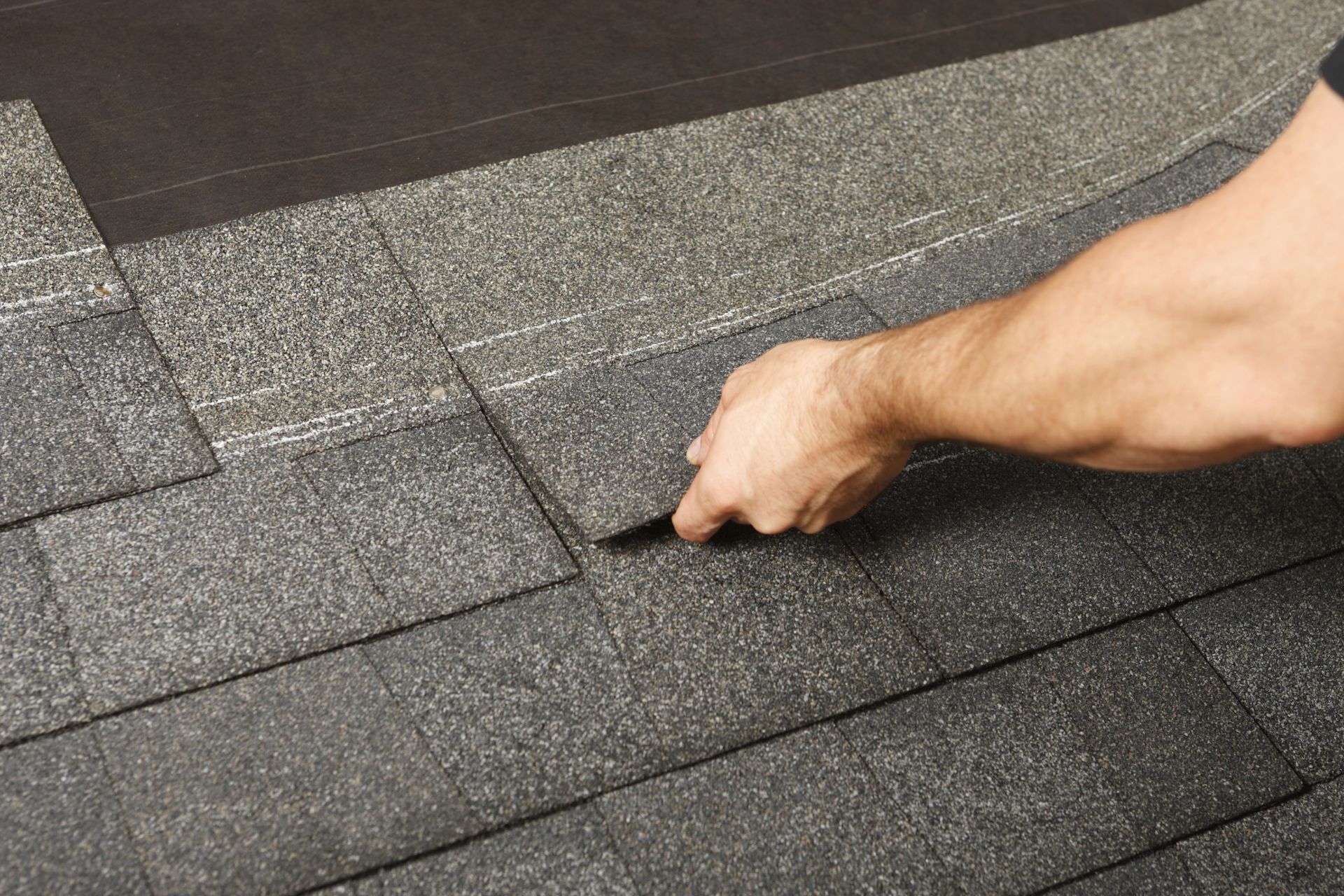 Hand inspecting and lifting a dark roof shingle on a gray shingled roof