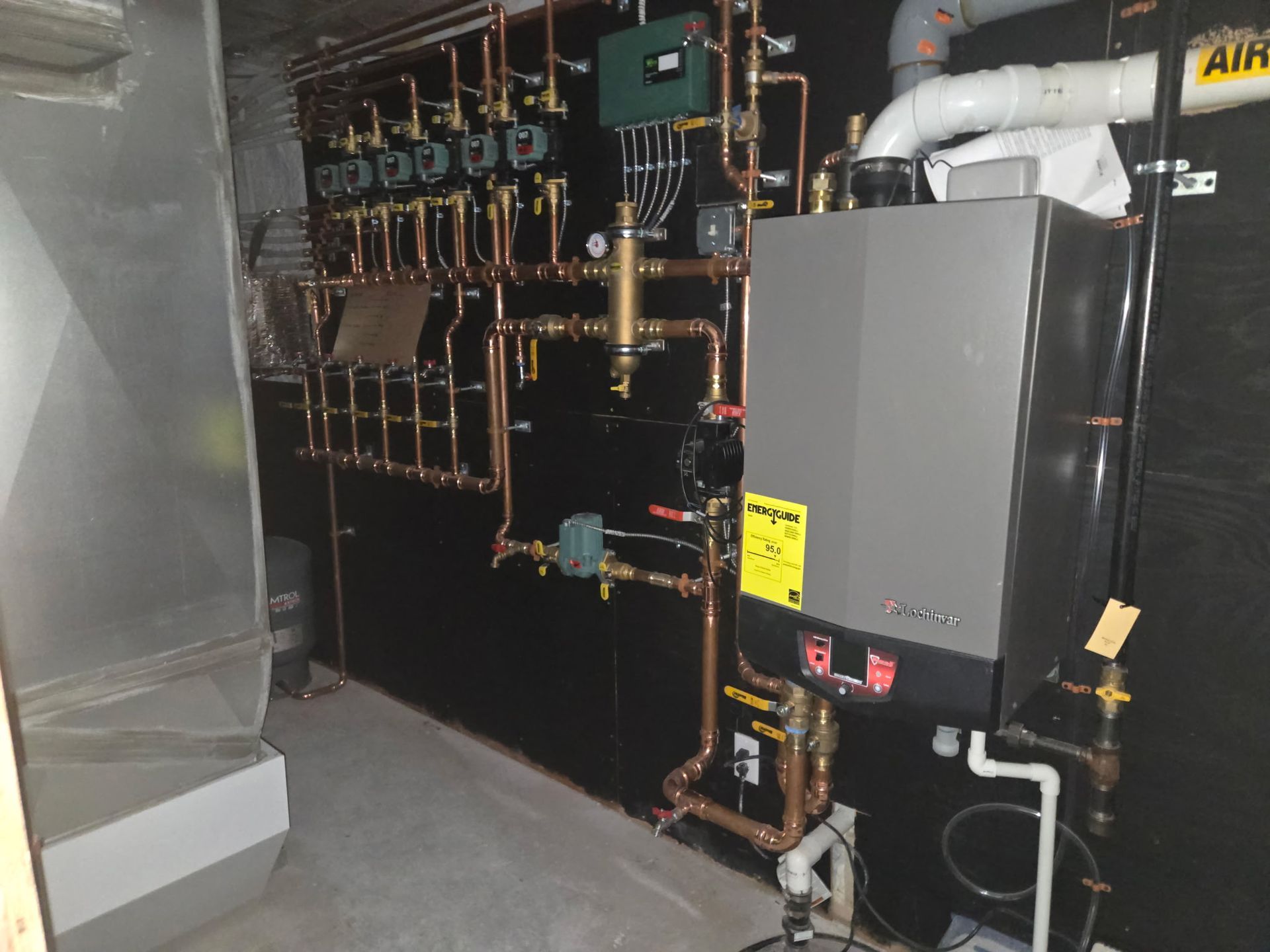 A gray wall-mounted boiler unit connected to a complex system of copper plumbing, pumps, and valves in a utility room.