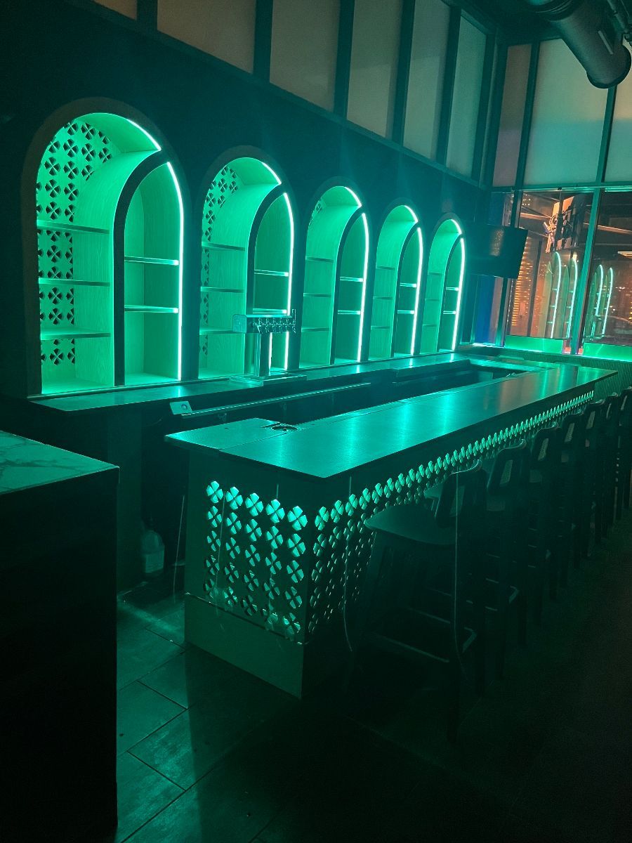 Green-lit bar with decorative archways and shelves. Empty bar counter in a dimly lit setting.