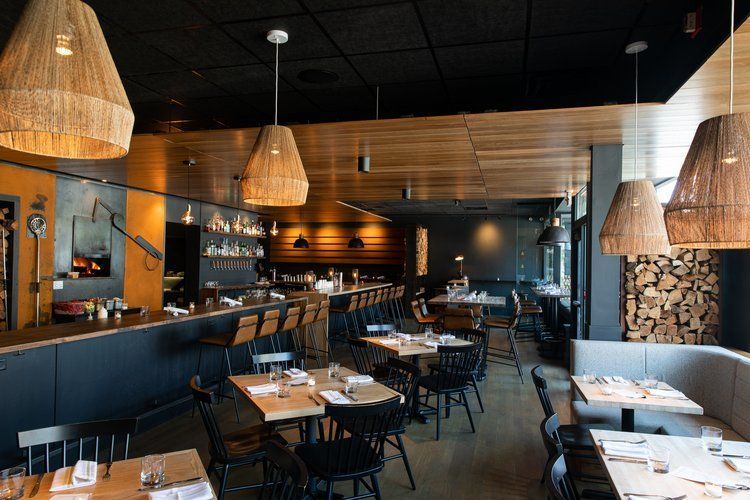 Restaurant interior: tables set, bar, woven pendant lights, wood accents, and a wood-fired oven.