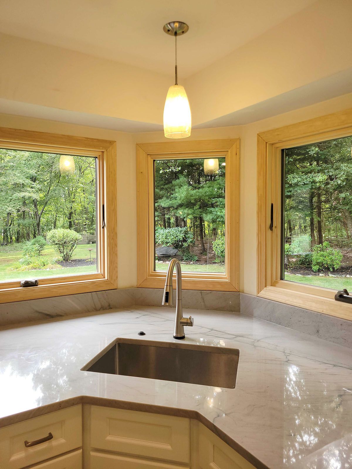 Kitchen corner with stainless steel sink, marble countertop, and windows overlooking a forest.