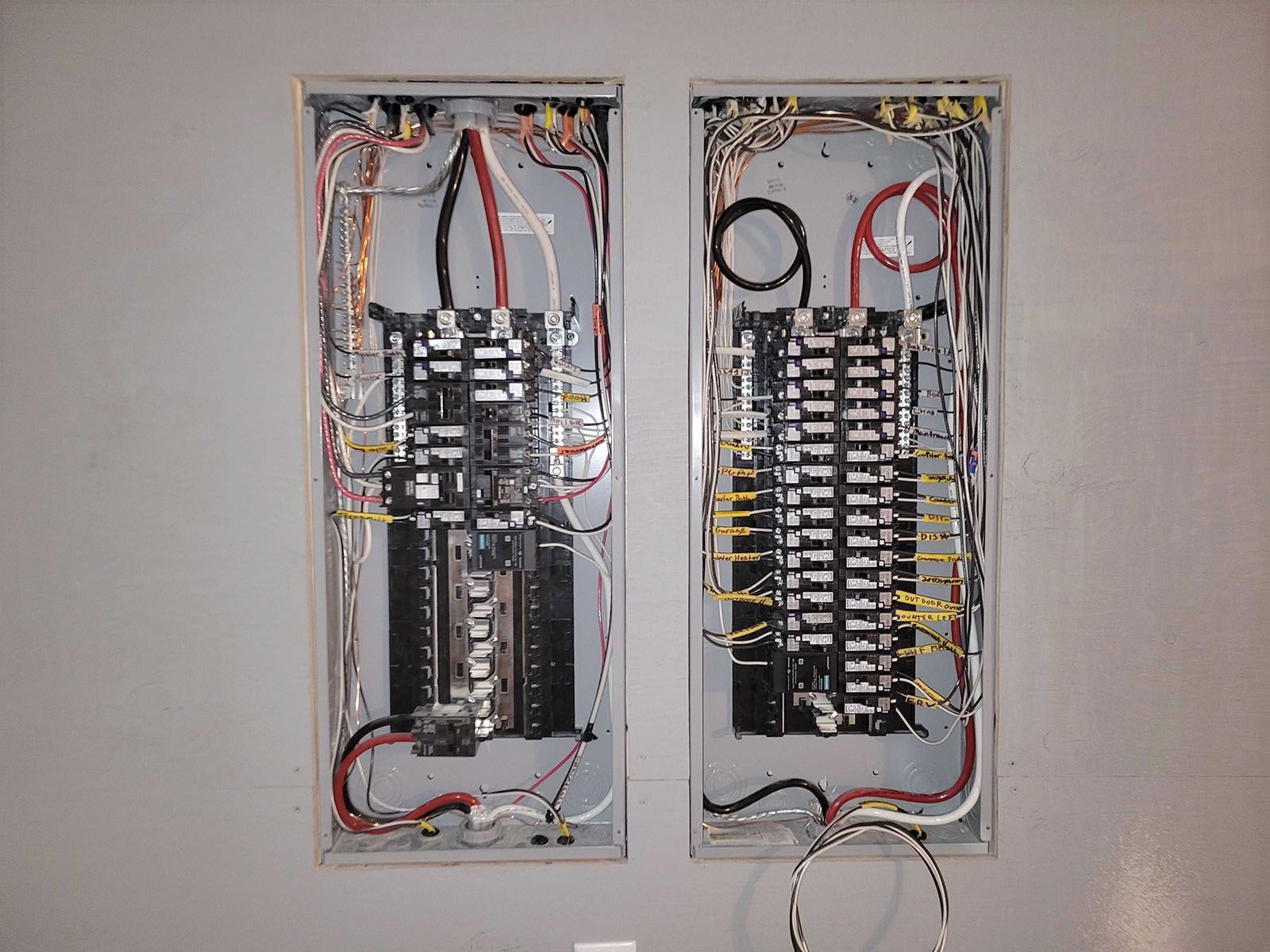 Two open electrical breaker panels with colored wires against a gray wall