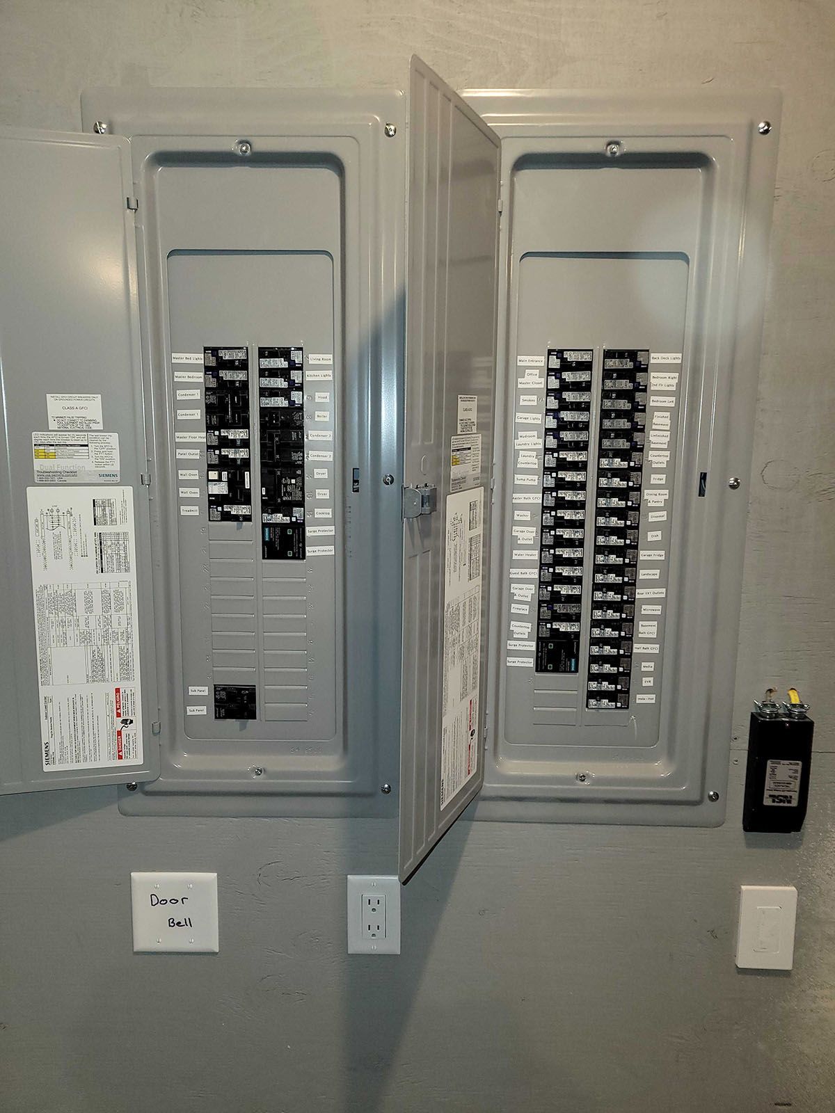 Two gray electrical panels, one open, mounted on a gray wall with an outlet and a switch nearby