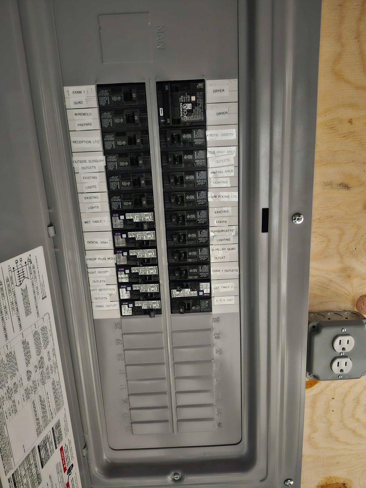 Open electrical panel with numerous circuit breakers, attached to a wooden surface