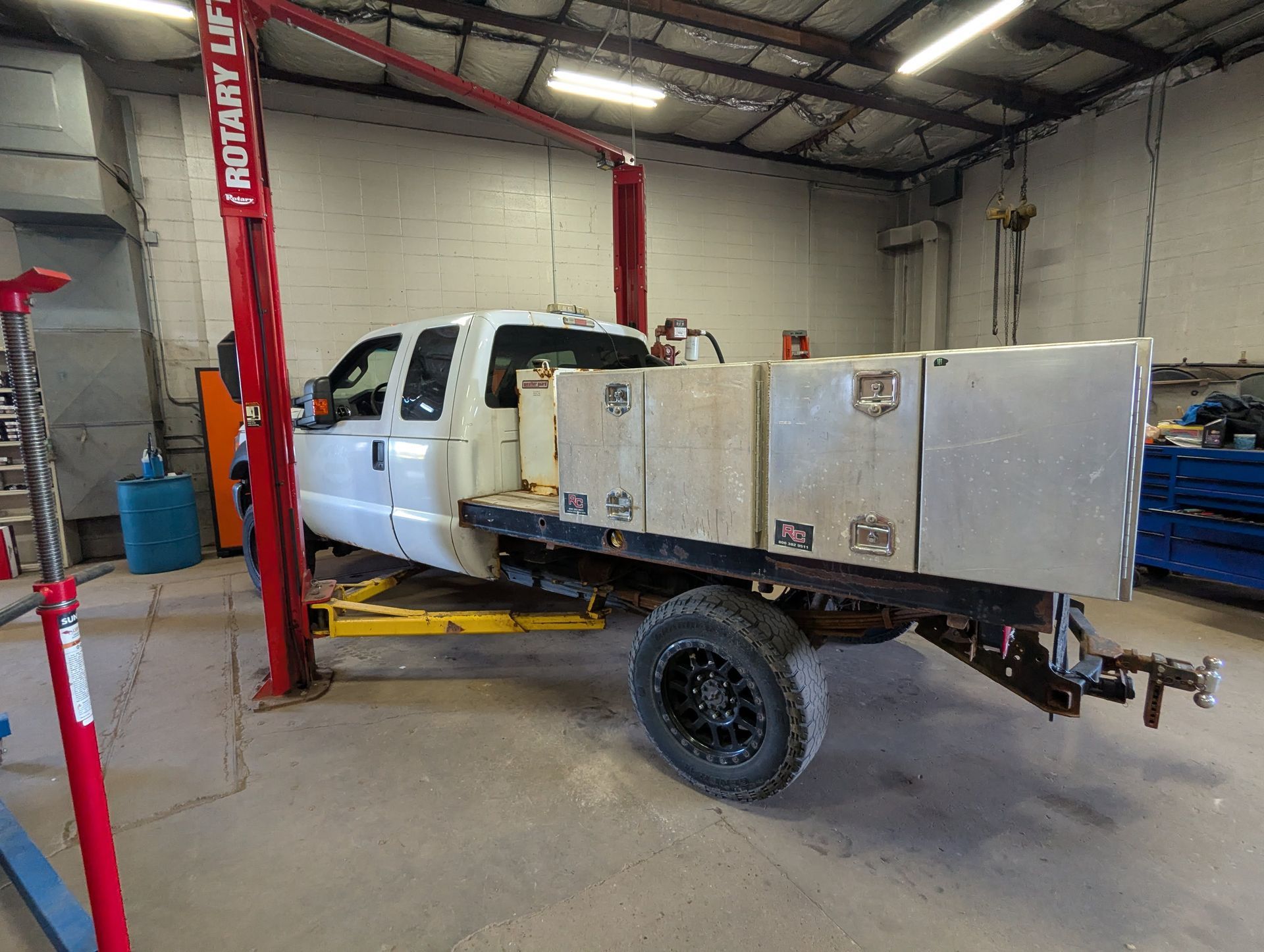 White pickup truck on a lift in an auto repair shop, with a large metal storage box in the bed.
