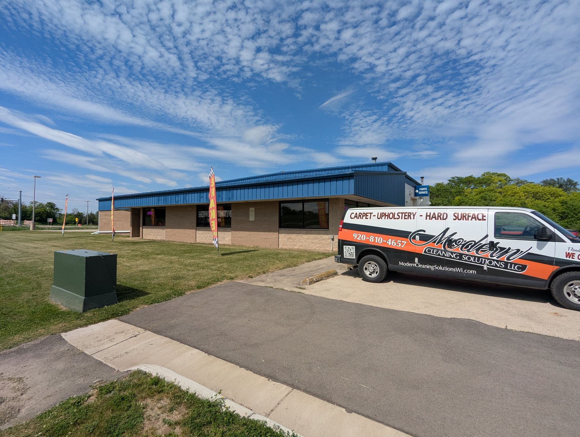 Low brick building with blue roof beside a white service van and grassy field under a cloudy sky