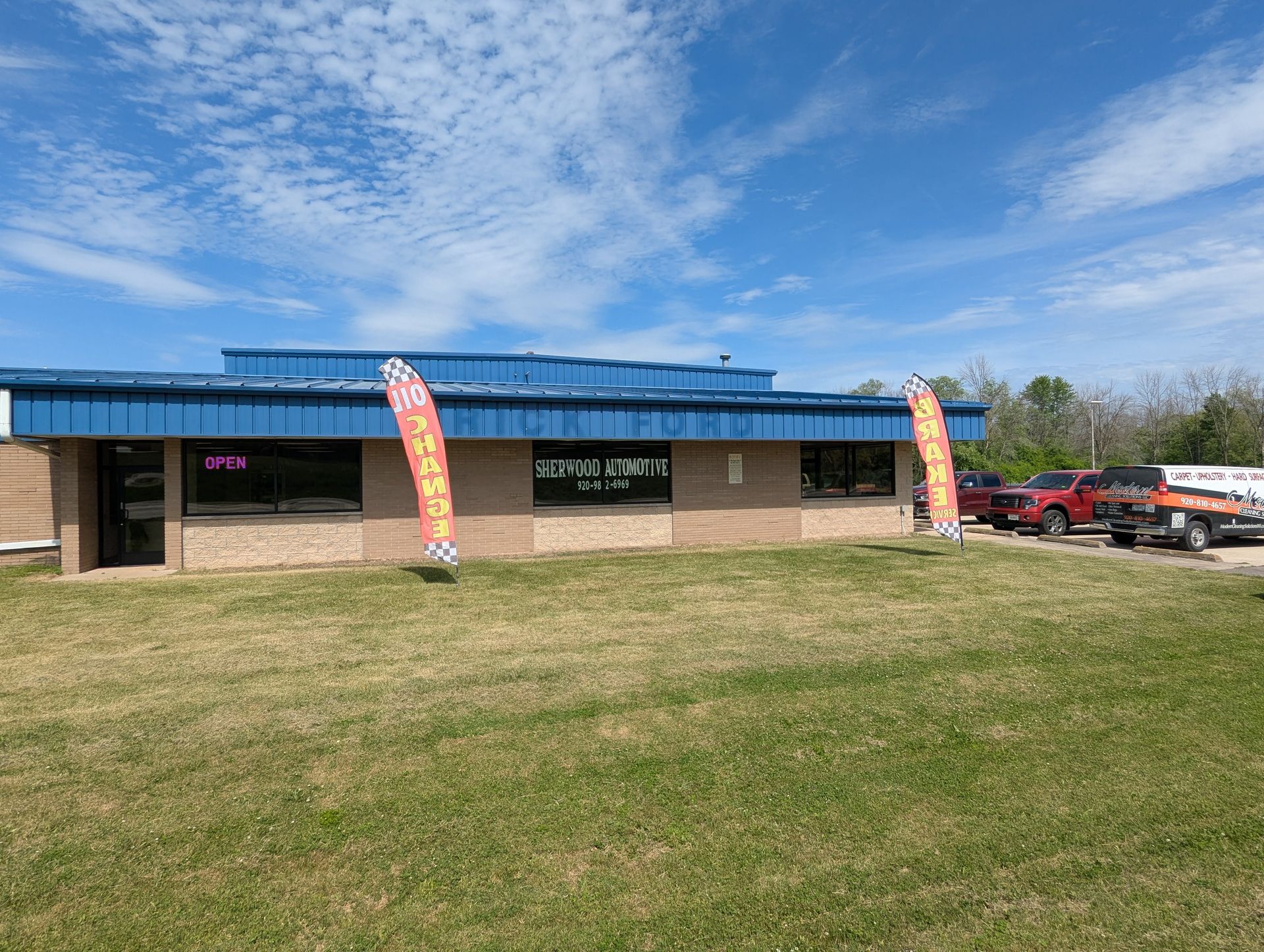 Single-story storefront with blue awning, grassy lot, and two orange flags under a partly cloudy sky
