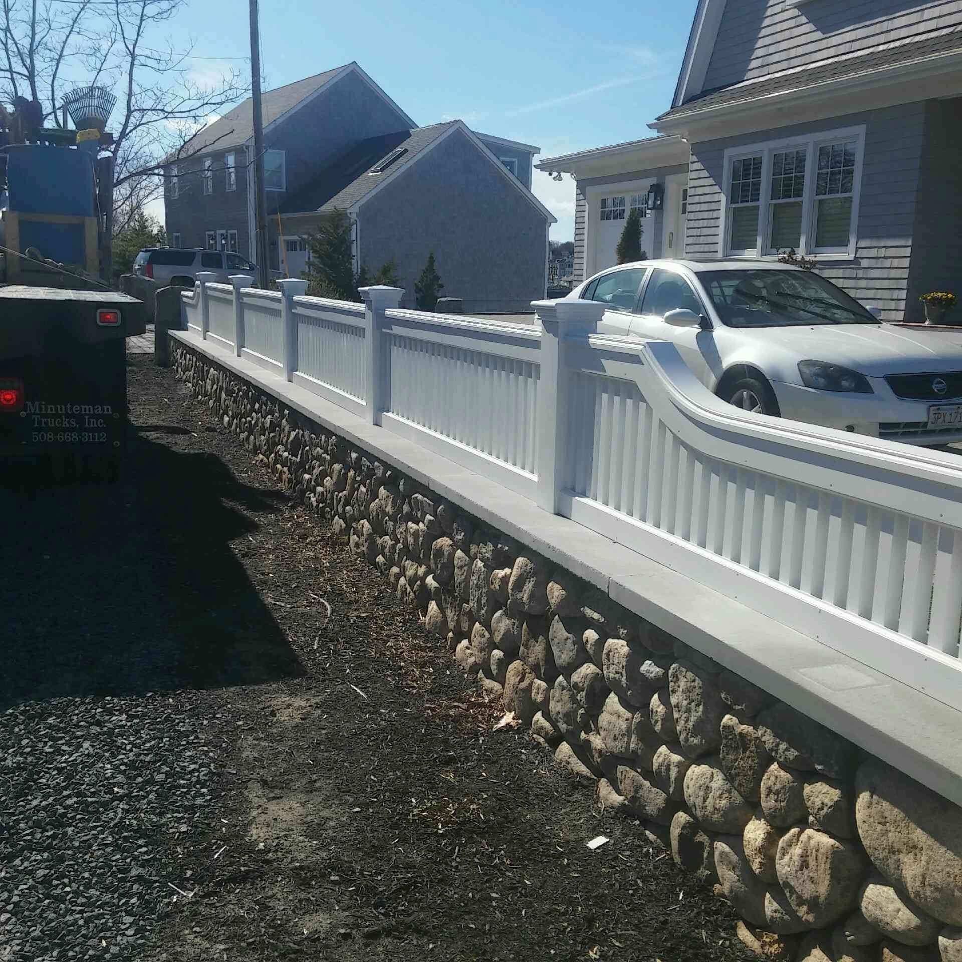 White picket fence on a stone wall beside a driveway, houses in the background.