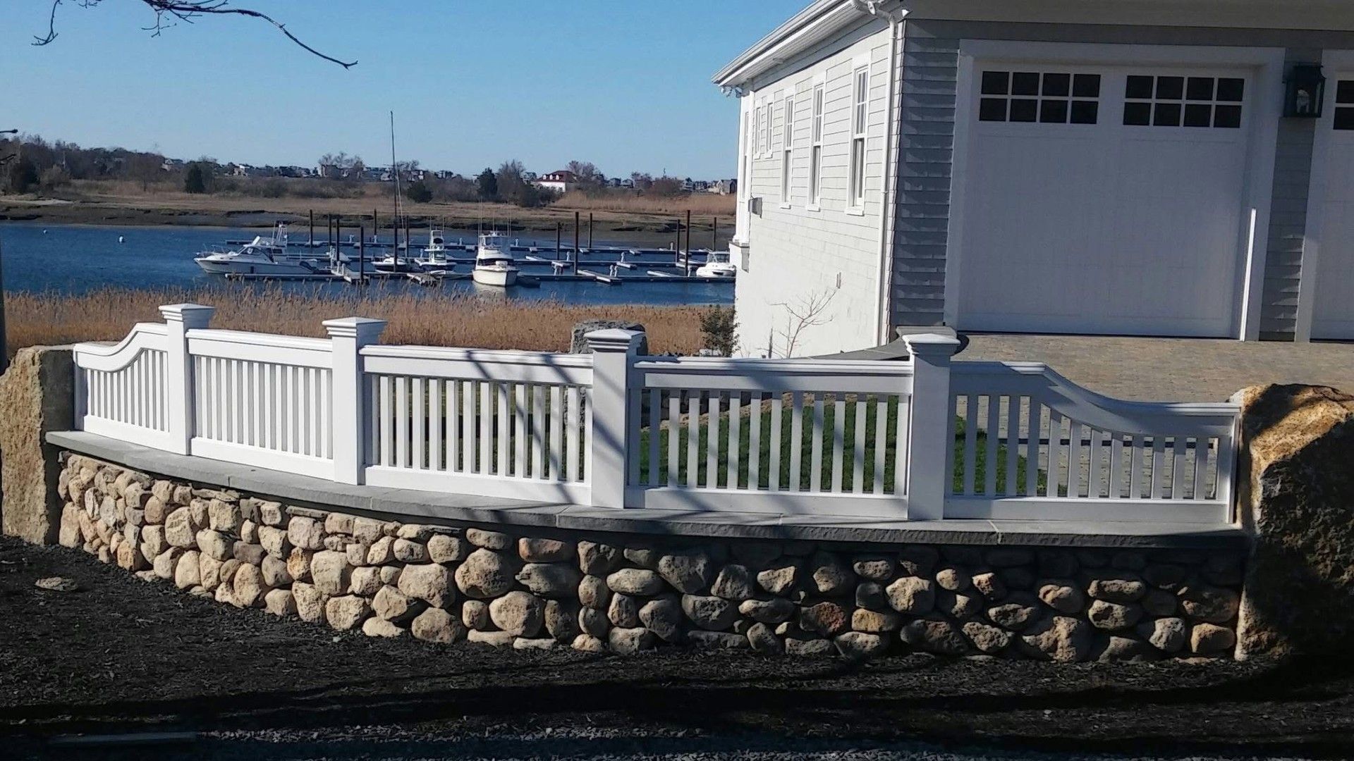 White picket fence on stone wall overlooking a harbor with boats, next to a white building.