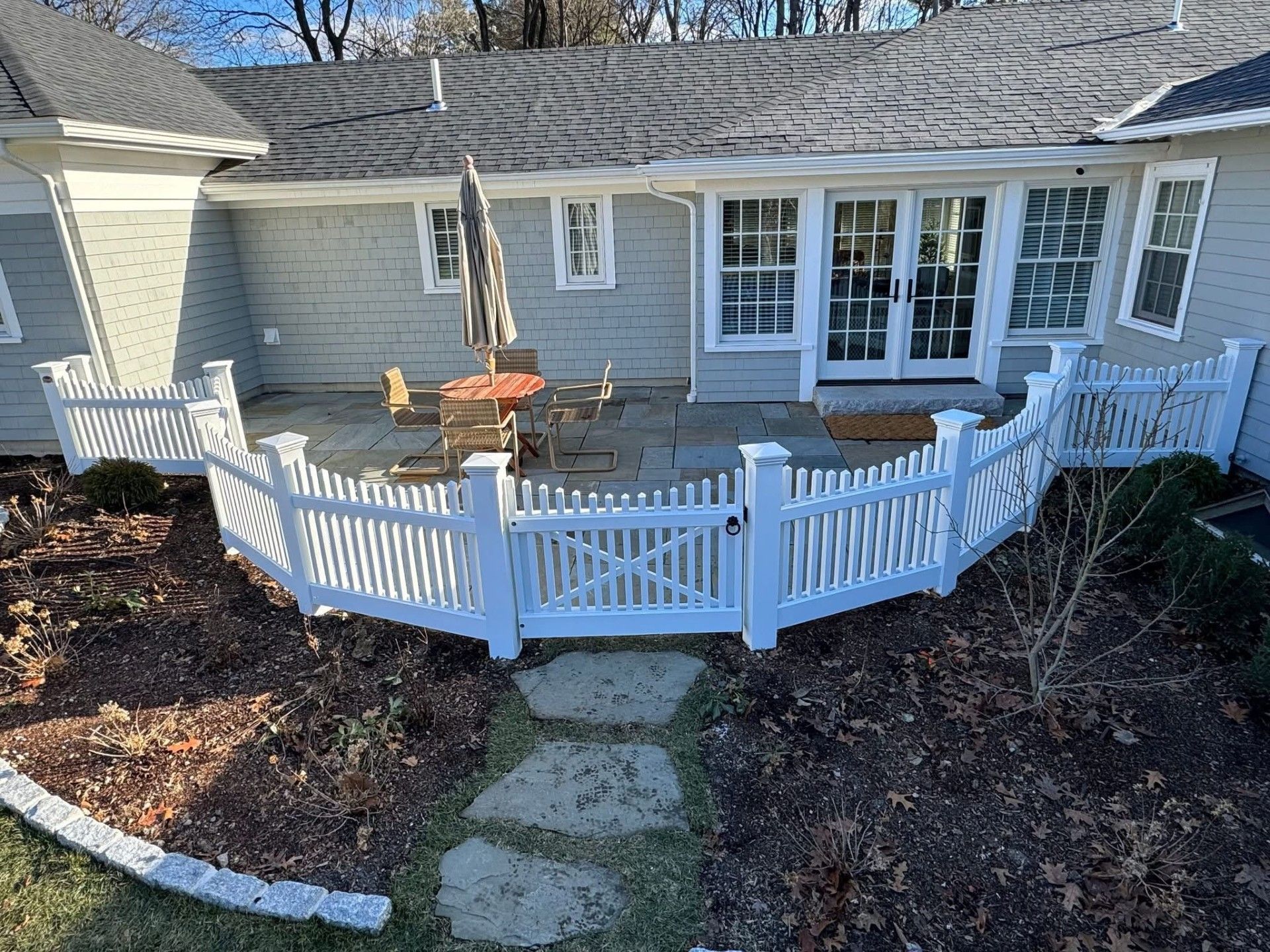 White picket fence encloses a patio with outdoor furniture, next to a light gray house with french doors.