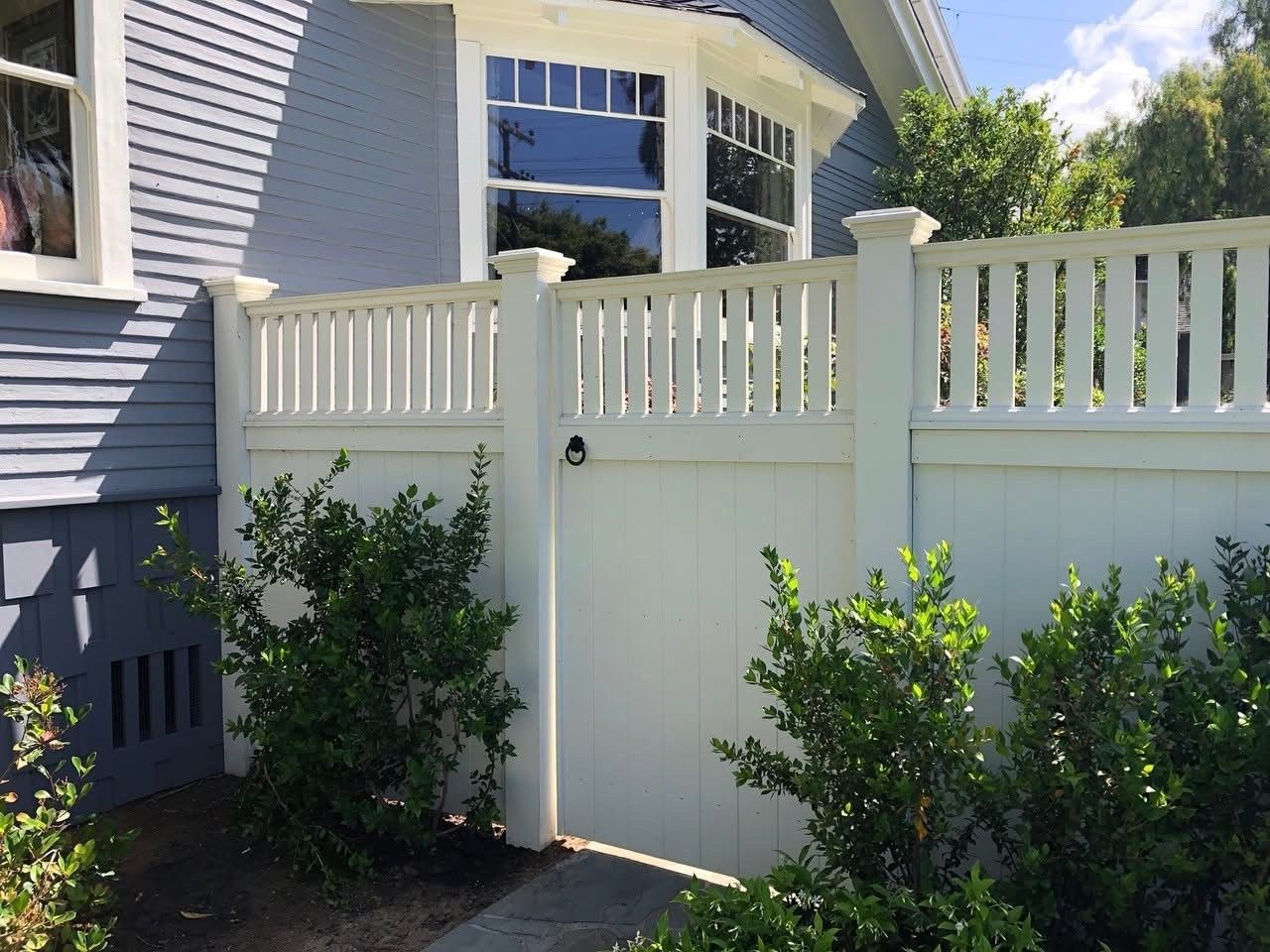 White fence with gate in front of a blue house with bay windows and green bushes.
