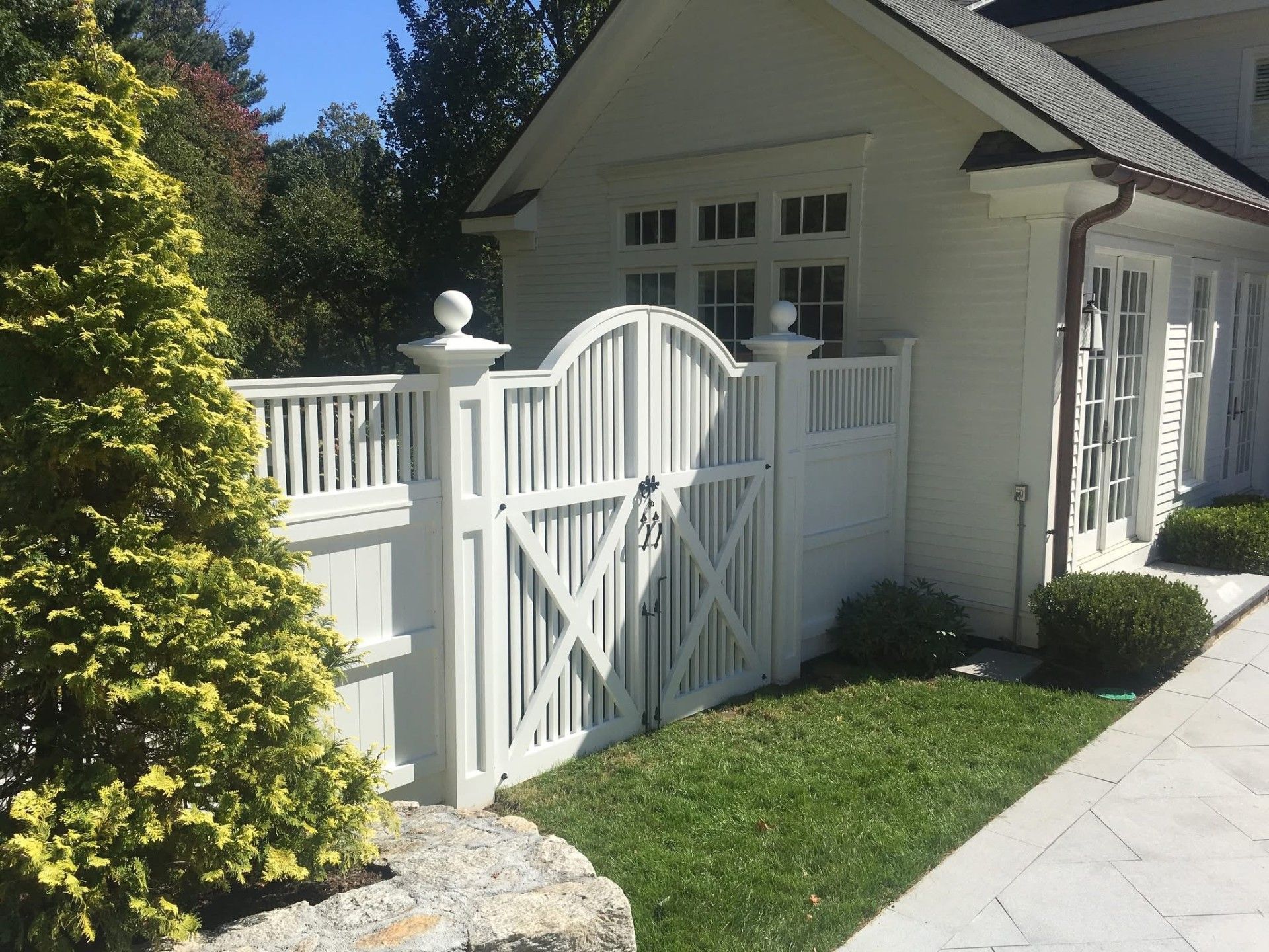 White picket fence with gate next to a white building. Lush green grass and small bushes.