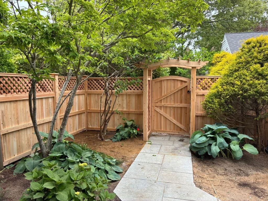 Wooden garden gate beside a stone path, surrounded by trees, a fence, and leafy plants.