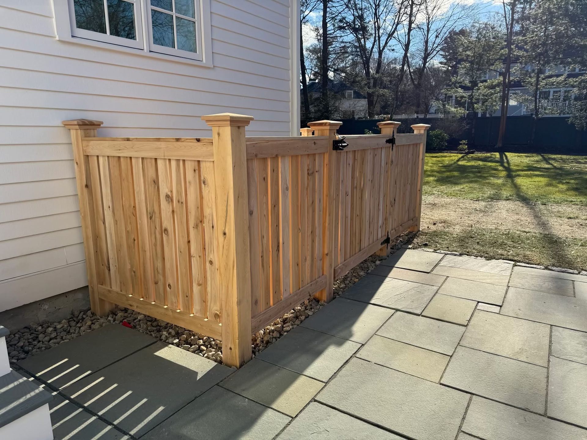Wooden privacy fence beside a house, bordering a paved patio and grassy yard.