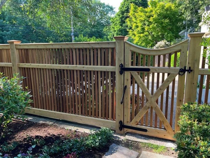 Wooden backyard fence with a tan gate and black hardware, bordered by greenery and trees.