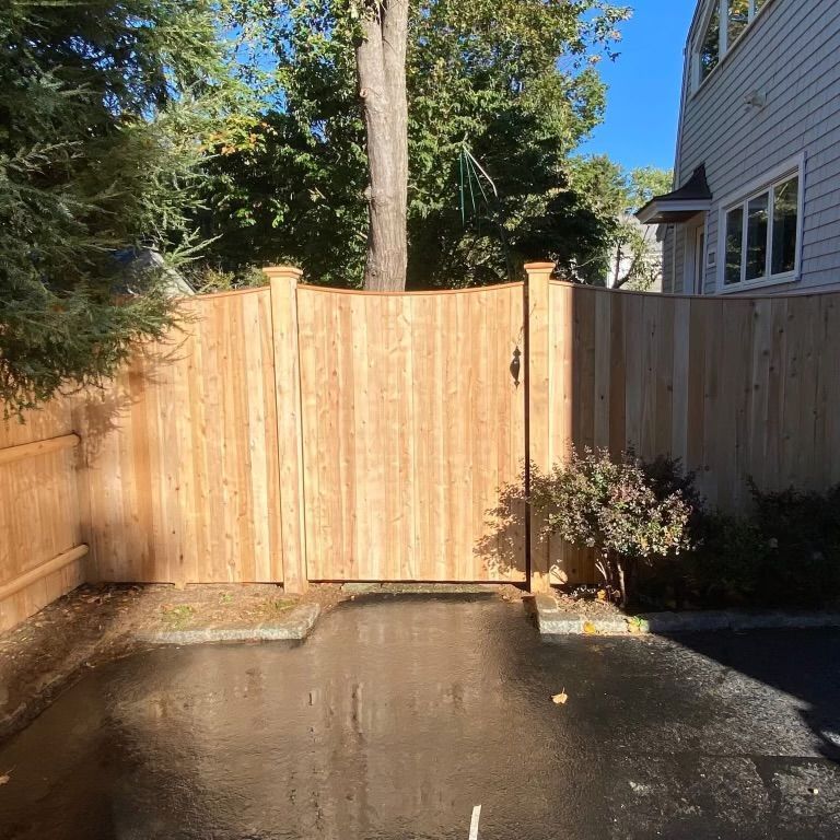 Wooden driveway gate between a house and fence, with wet pavement in front and trees overhead.