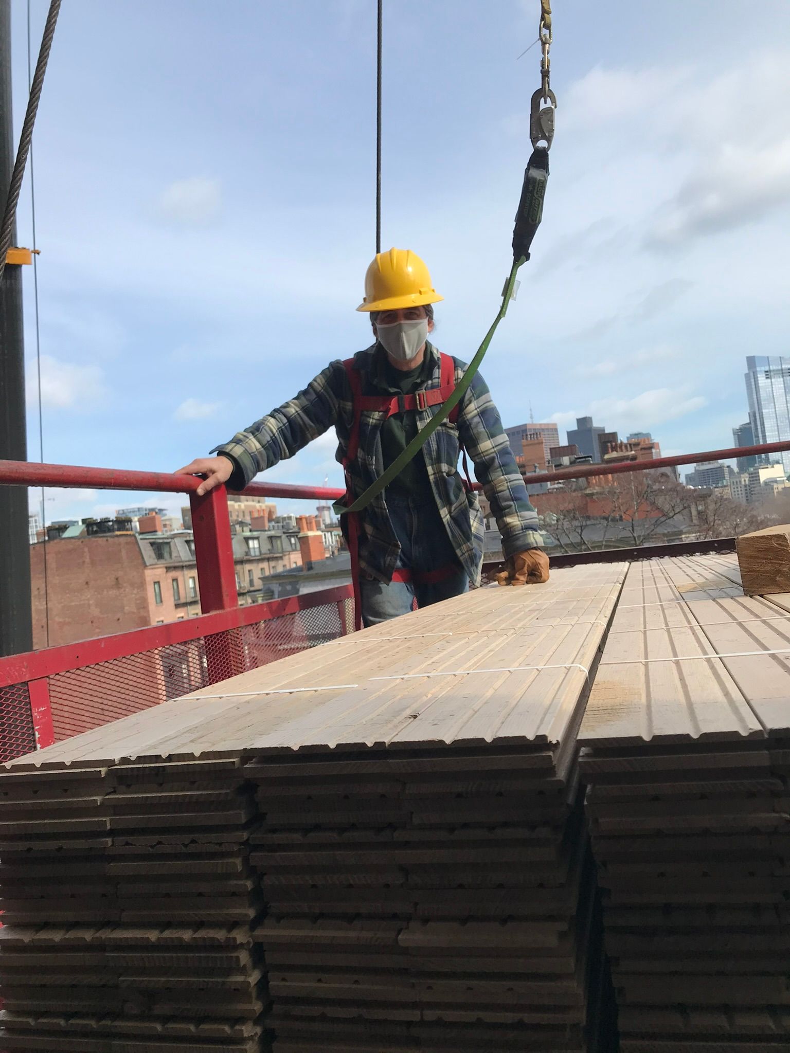 Construction worker on a platform, wearing a hard hat, mask, and harness, near a stack of wood planks, with a cityscape background.
