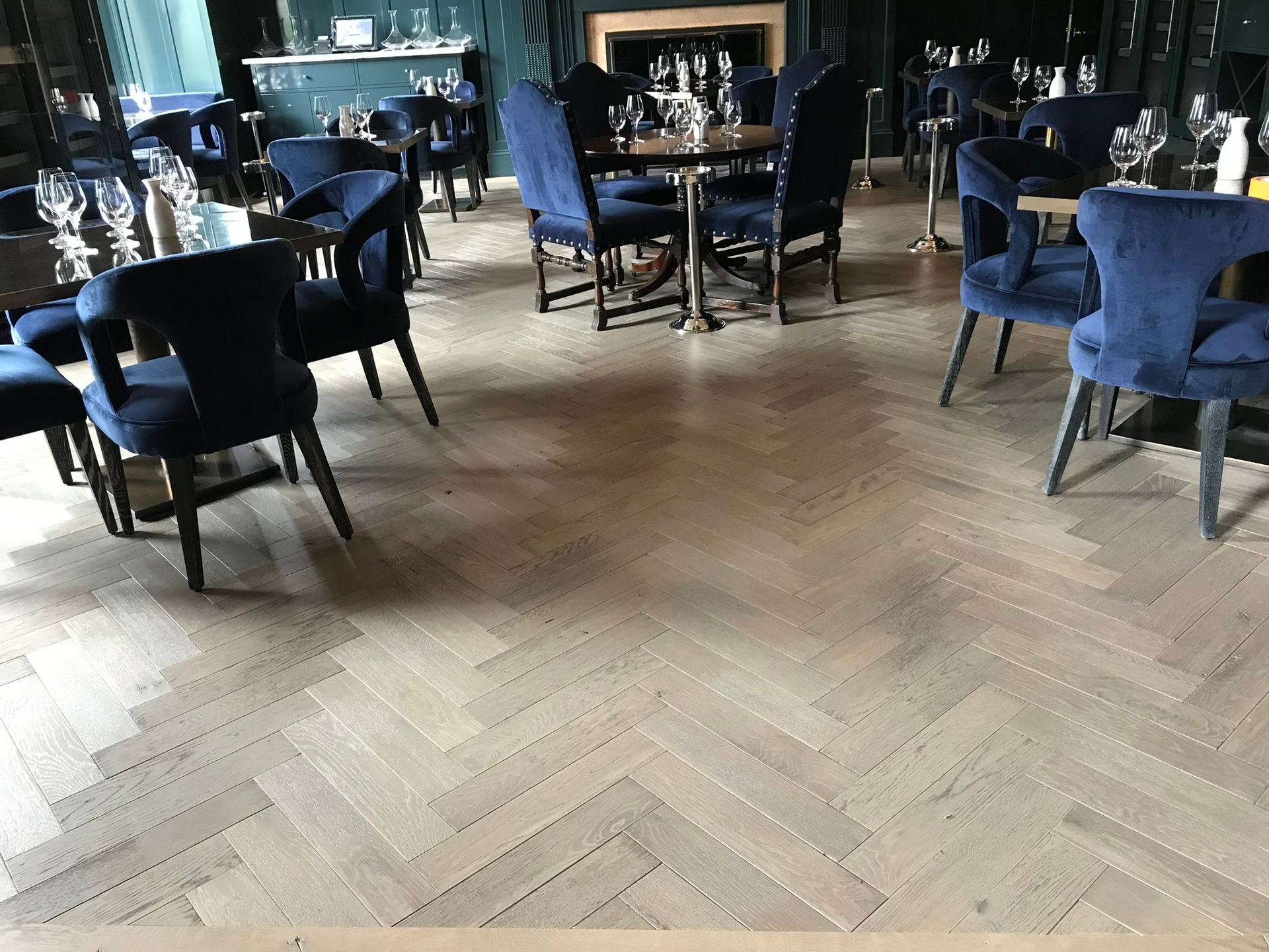 Restaurant interior with light wood parquet floor, blue velvet chairs, and round tables set with glasses.