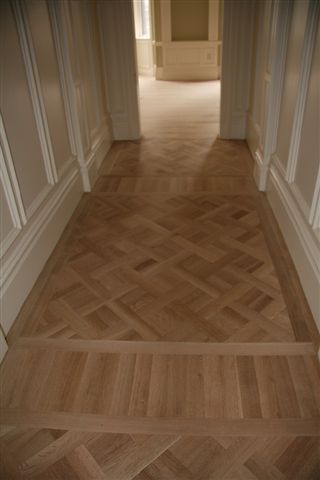 Hallway with parquet wood flooring in a decorative pattern, bordered by white walls and molding.