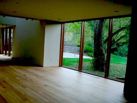 Empty room with large windows overlooking a green outdoor space, with wooden floors and trim.
