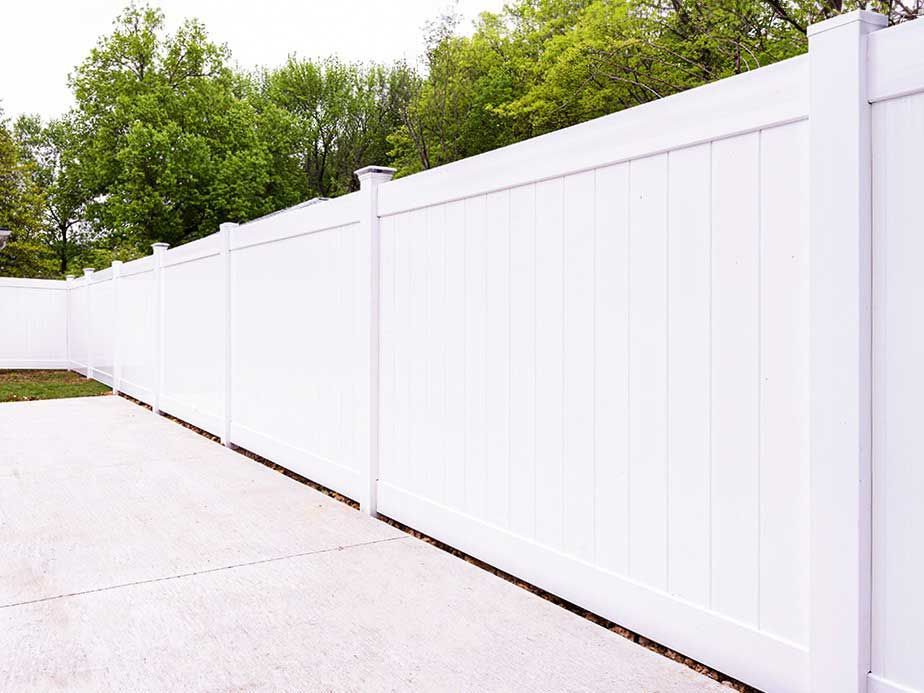 A white vinyl fence surrounds a driveway with trees in the background.