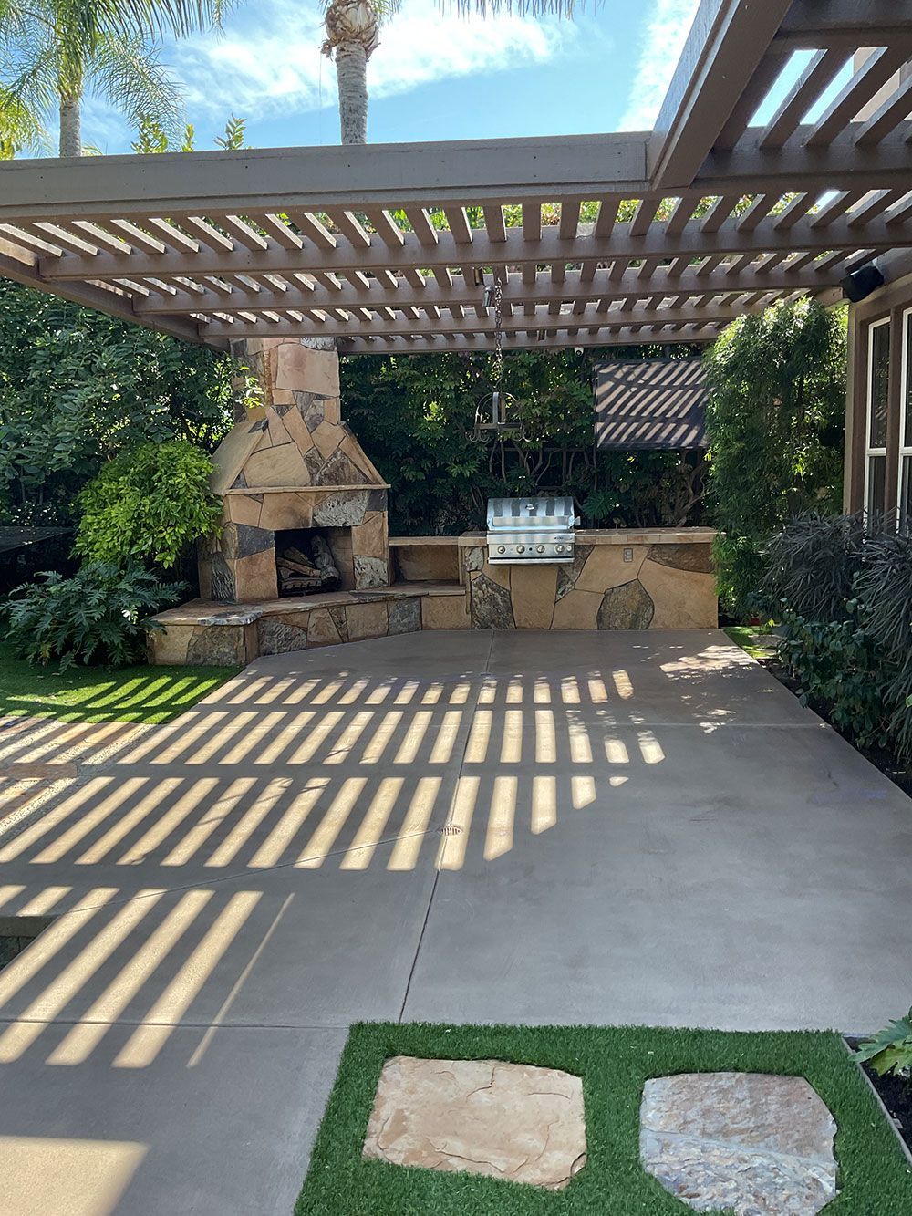 Outdoor patio with stone fireplace, grill, and pergola casting shadows on concrete flooring.