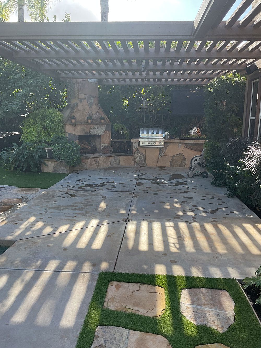Outdoor patio with pergola, fireplace, and grill, over concrete and stone pavers, greenery in background.