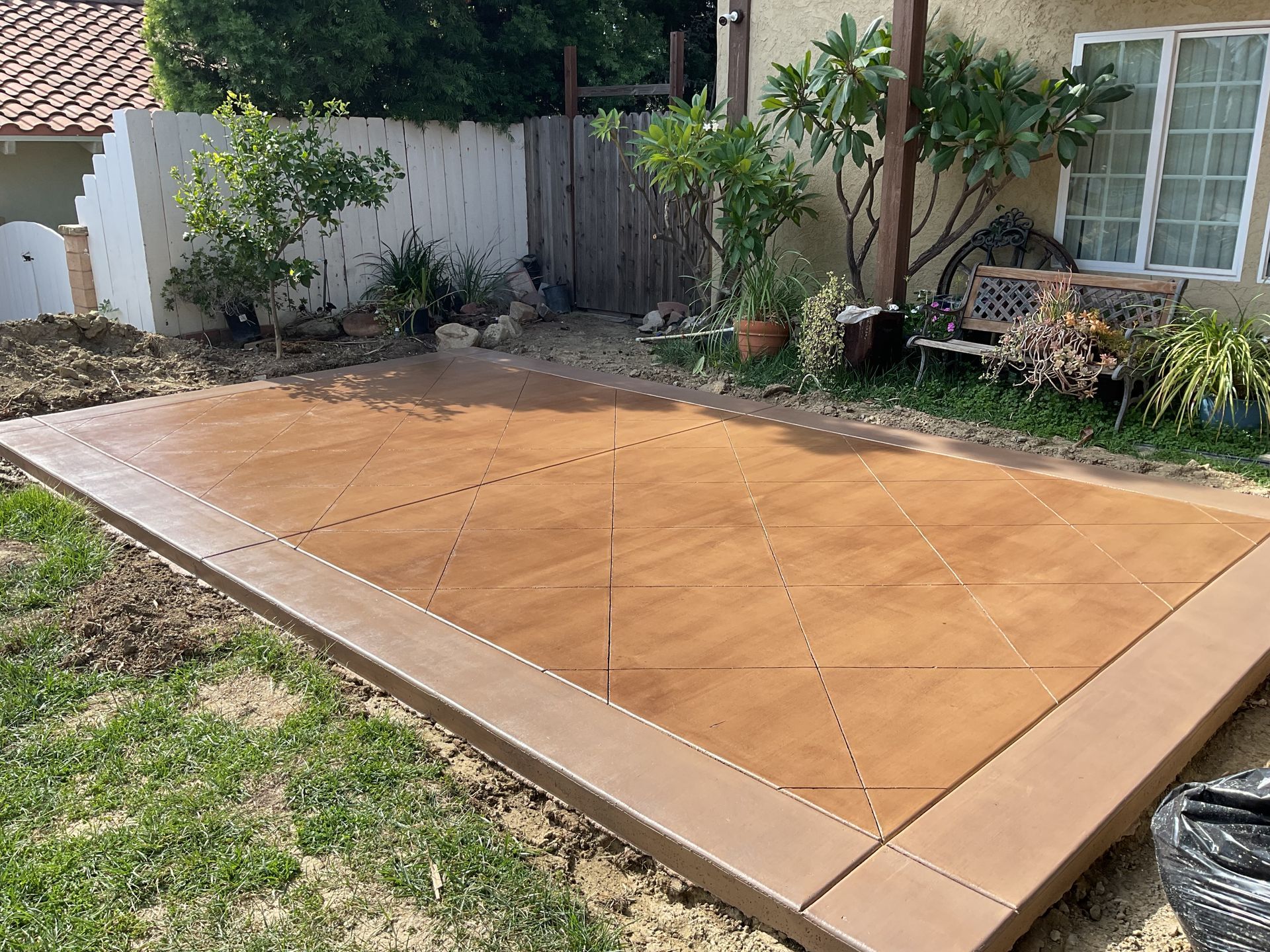 Freshly poured tan concrete patio in a backyard, bordered by a house, fence, and garden plants.