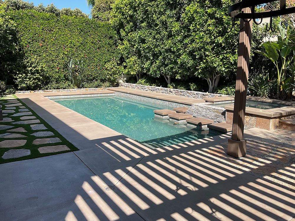 Swimming pool surrounded by concrete patio and greenery, with a pergola casting shadows.