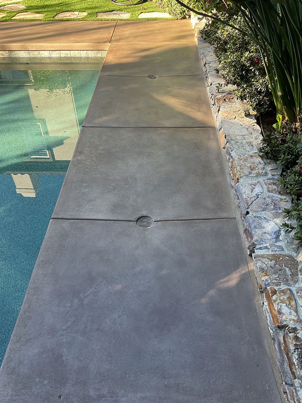 Poolside concrete border with dark gray surface, next to turquoise pool and brick wall with plants.