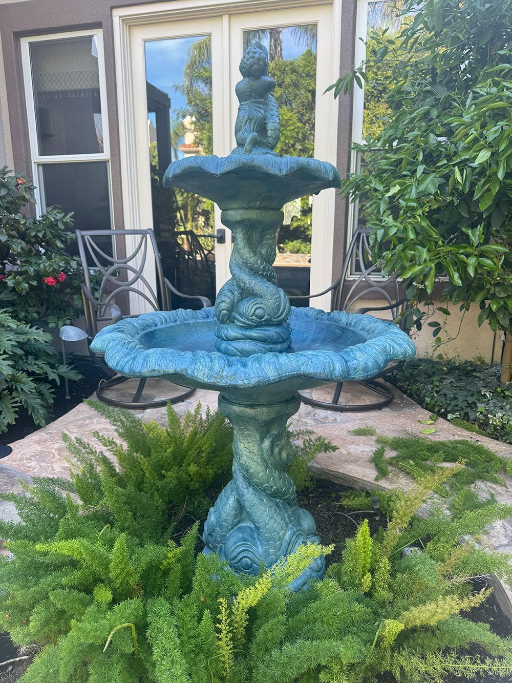Blue tiered outdoor water fountain surrounded by green ferns, in front of a glass door.