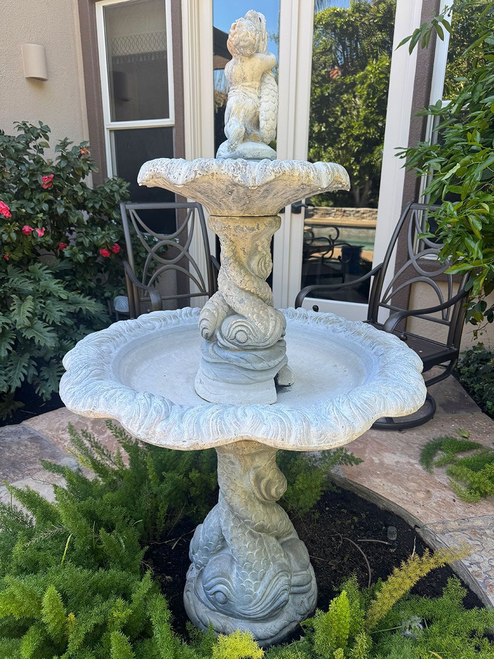Stone water fountain with cherub statue, surrounded by greenery in a yard.